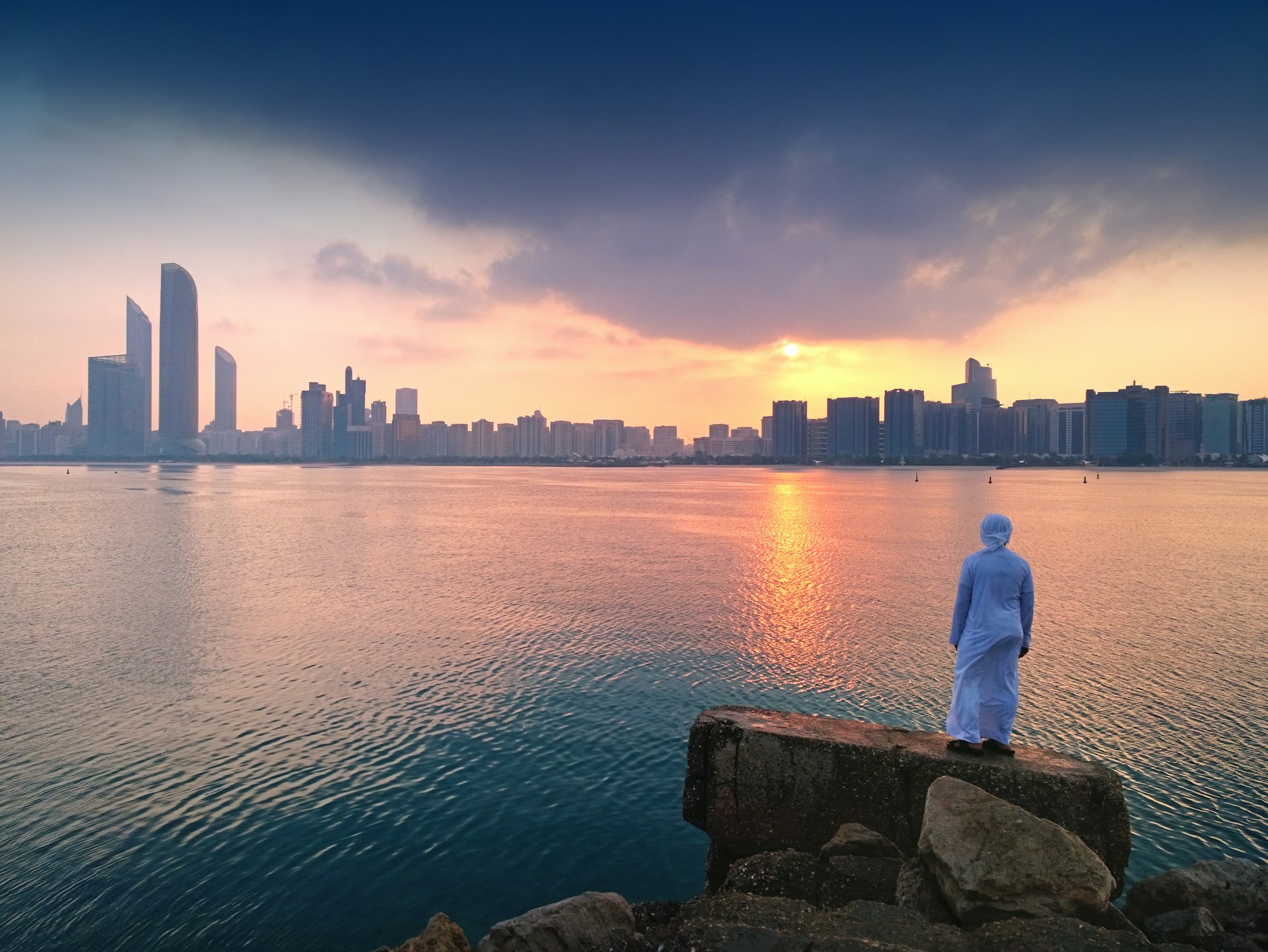 A person stands on a shoreline looking out towards the skyscraper's of Abu Dhabi's skyline