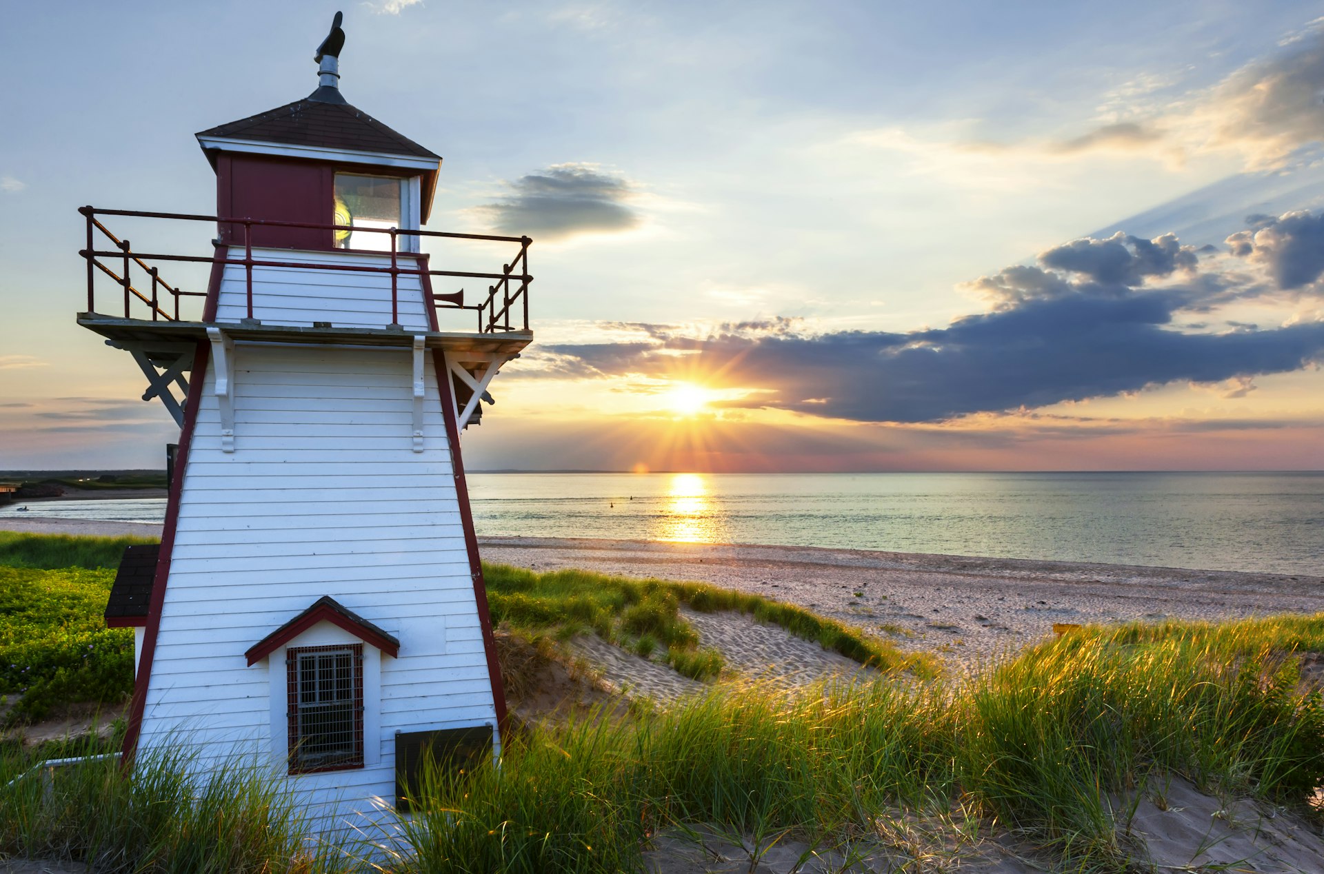A wooden lighthouse painted bright white stands in the foreground, as the sun sets over a calm sea beyond.