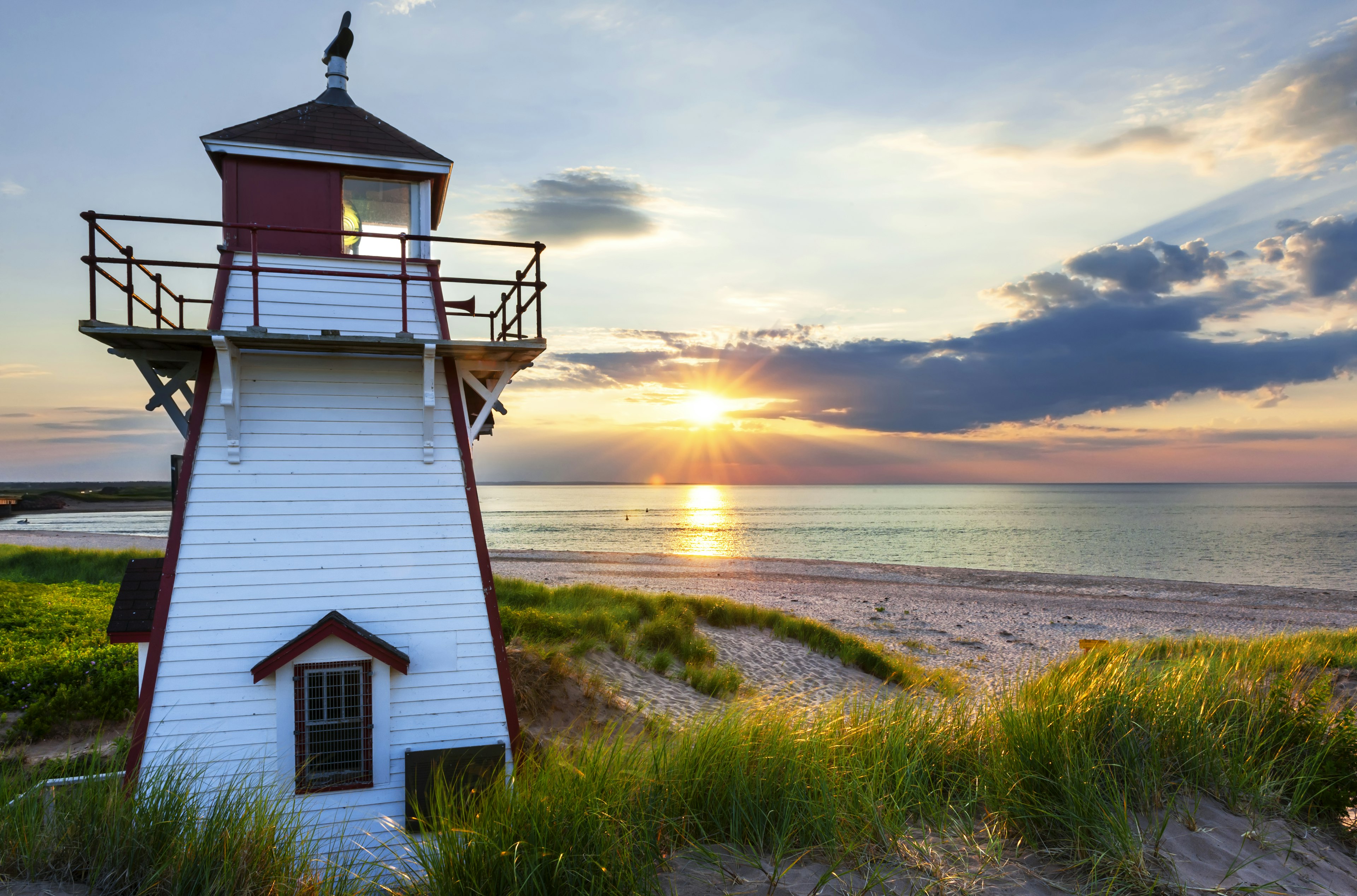 A wooden lighthouse painted bright white stands in the foreground, as the sun sets over a calm sea beyond.