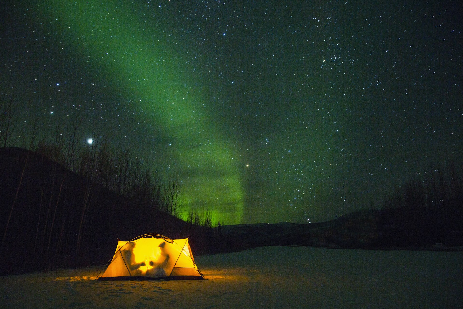 A warmly lit tent on a snowy ground at night with the green hues of the Northern Lights in the background, under a star-filled sky.