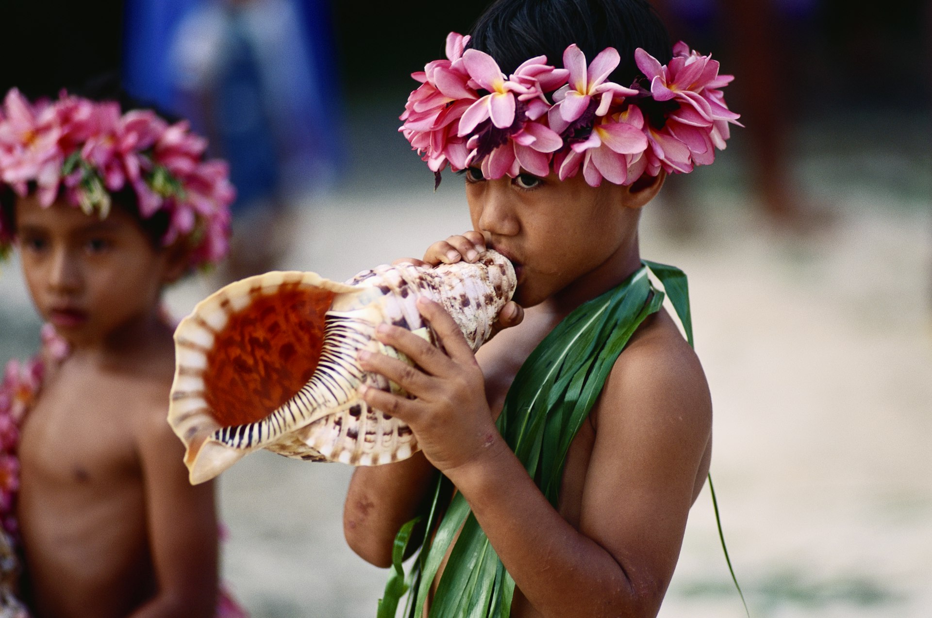 A child wearing a flower headdress blows on a conch horn