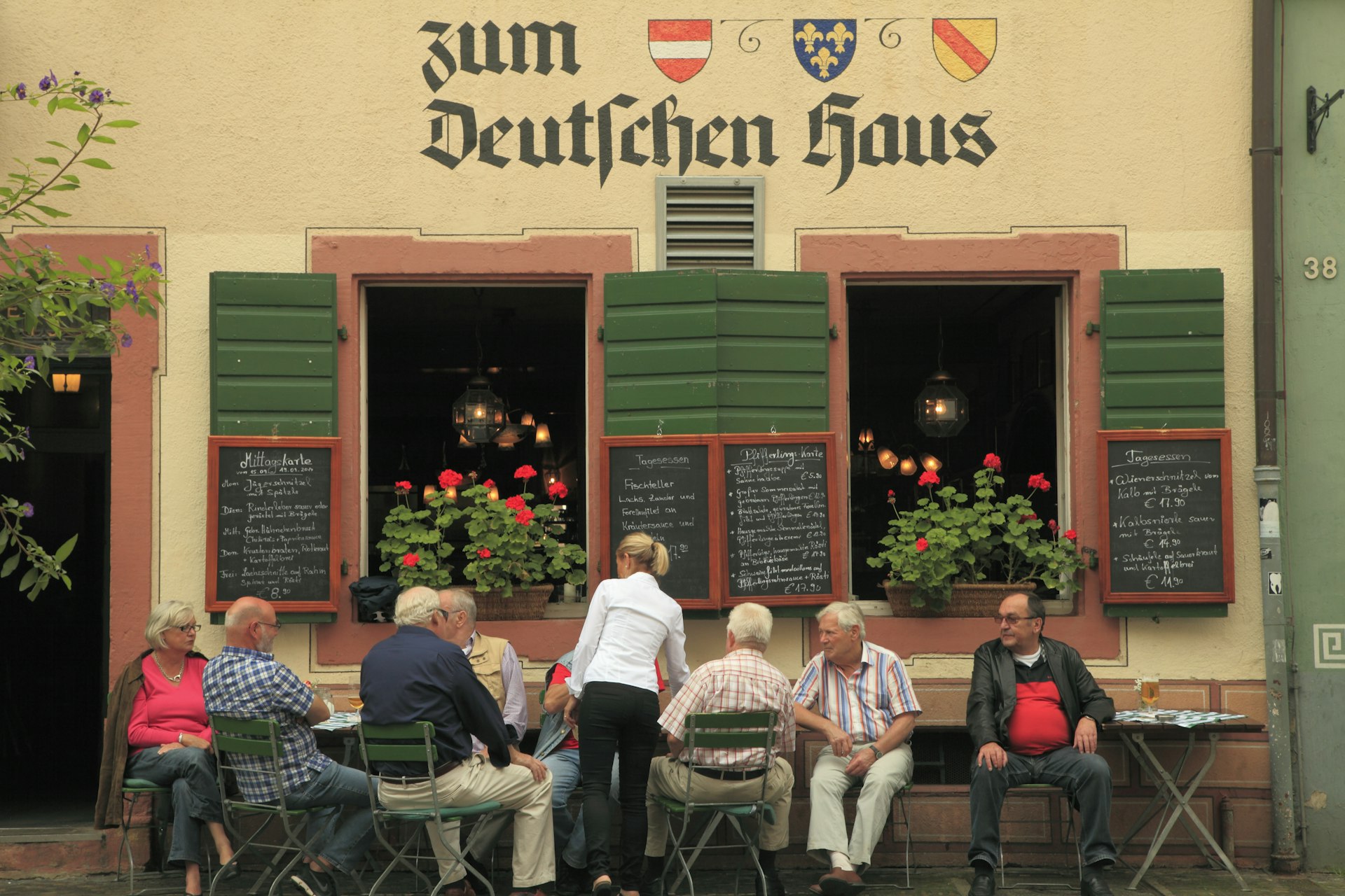 Patrons seated outside a traditional German restaurant with a sign reading 'Zum Deutschen Haus', menus displayed on a blackboard, and an exterior adorned with flowers.