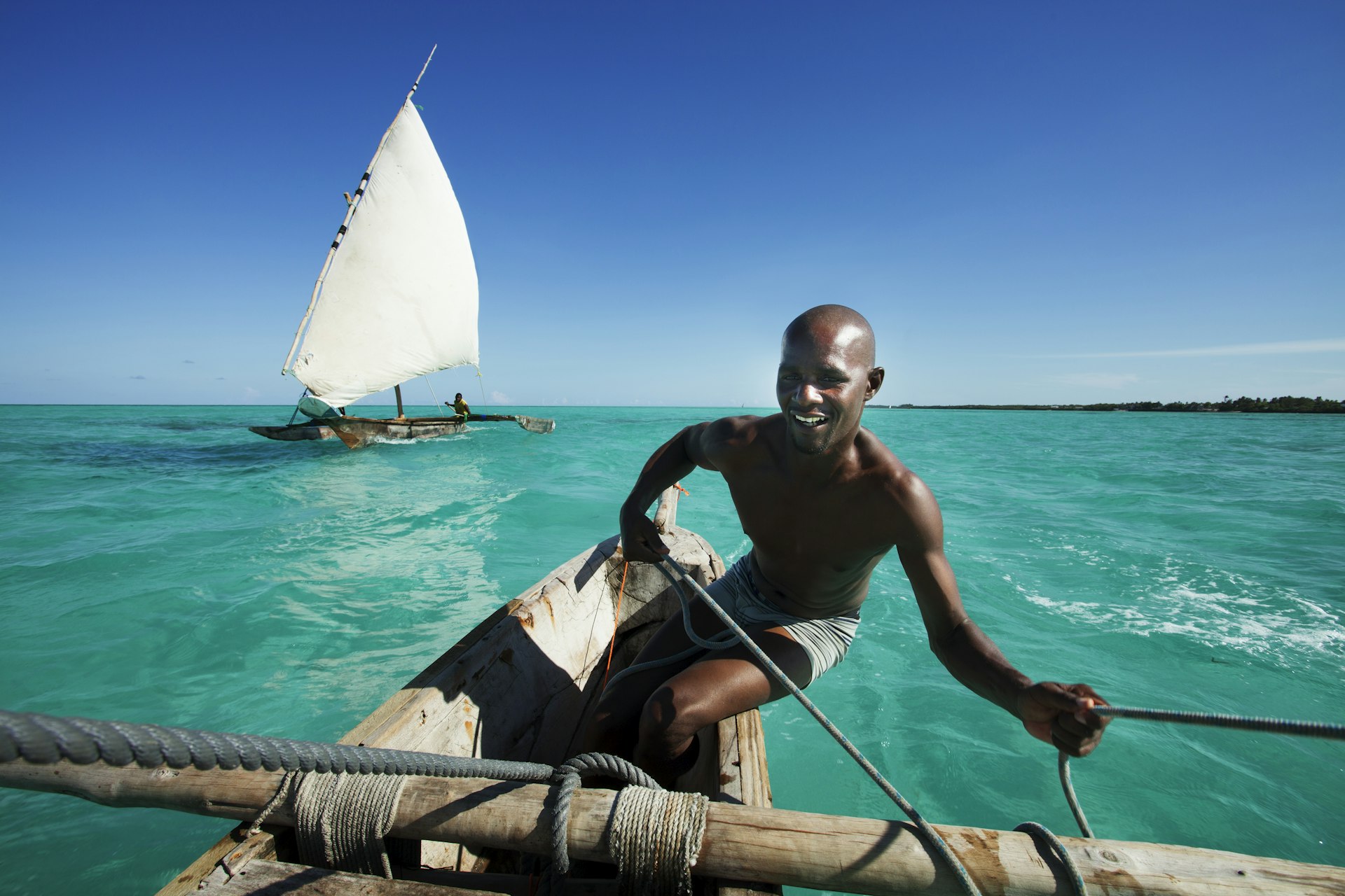 A traditional dhow sailing boat with a triangular sail on turquoise waters with a clear sky, manned by a smiling sailor leaning on the wooden hull.