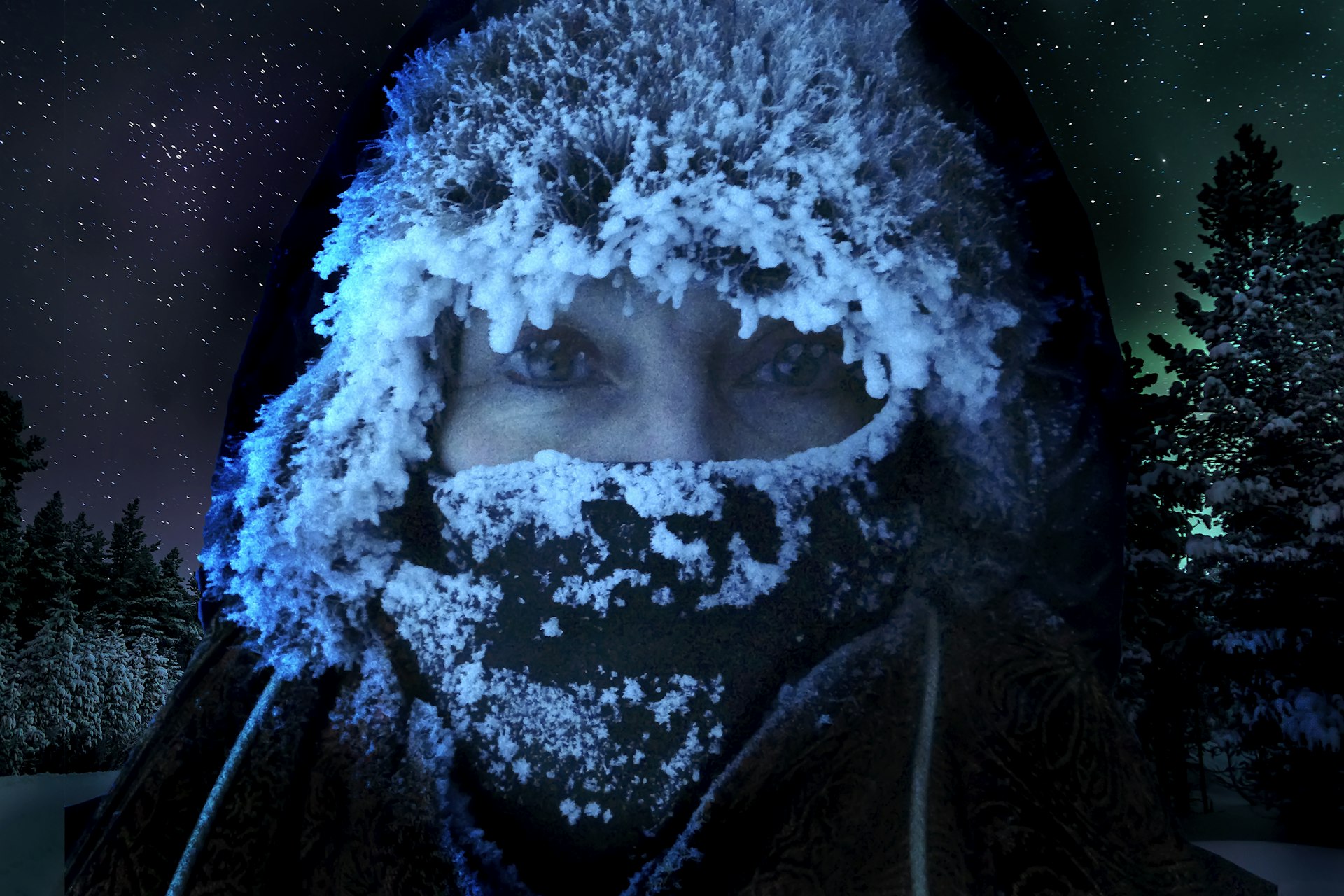 Close-up of a person's face covered with frost and ice on their hood and scarf, with the Northern Lights visible in the night sky behind them and trees faintly visible in the darkness