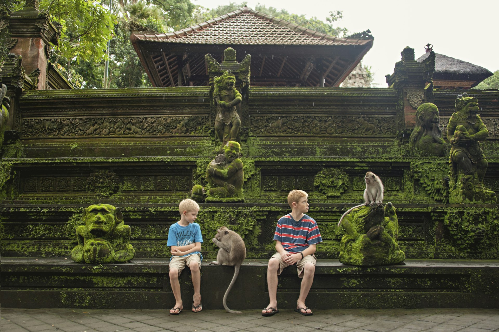 Two boys sit on the wall outside a temple looking at two monkeys