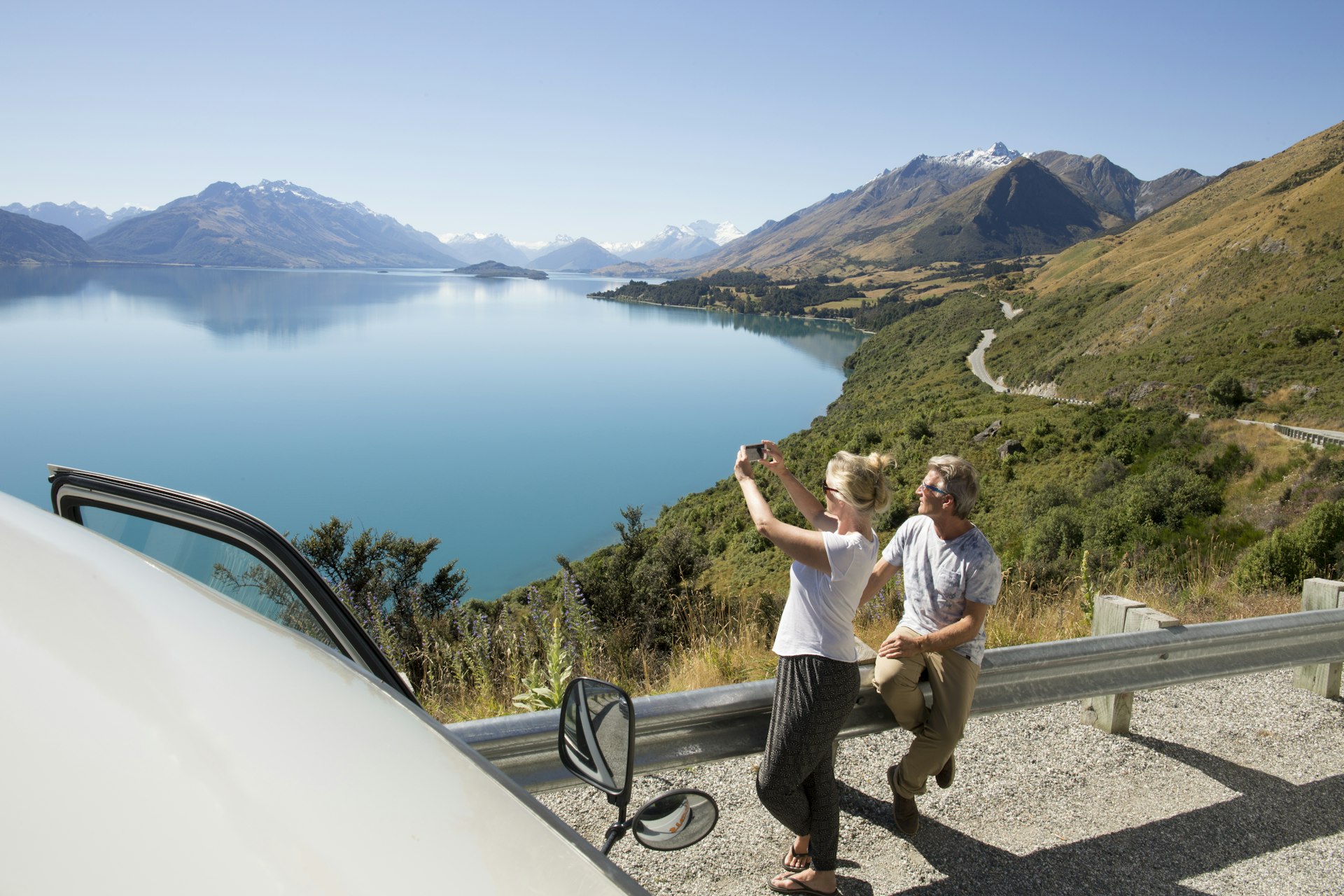 Woman takes pic beside camper van overlooking lake and mountain in Queenstown, New Zealand while man relaxes