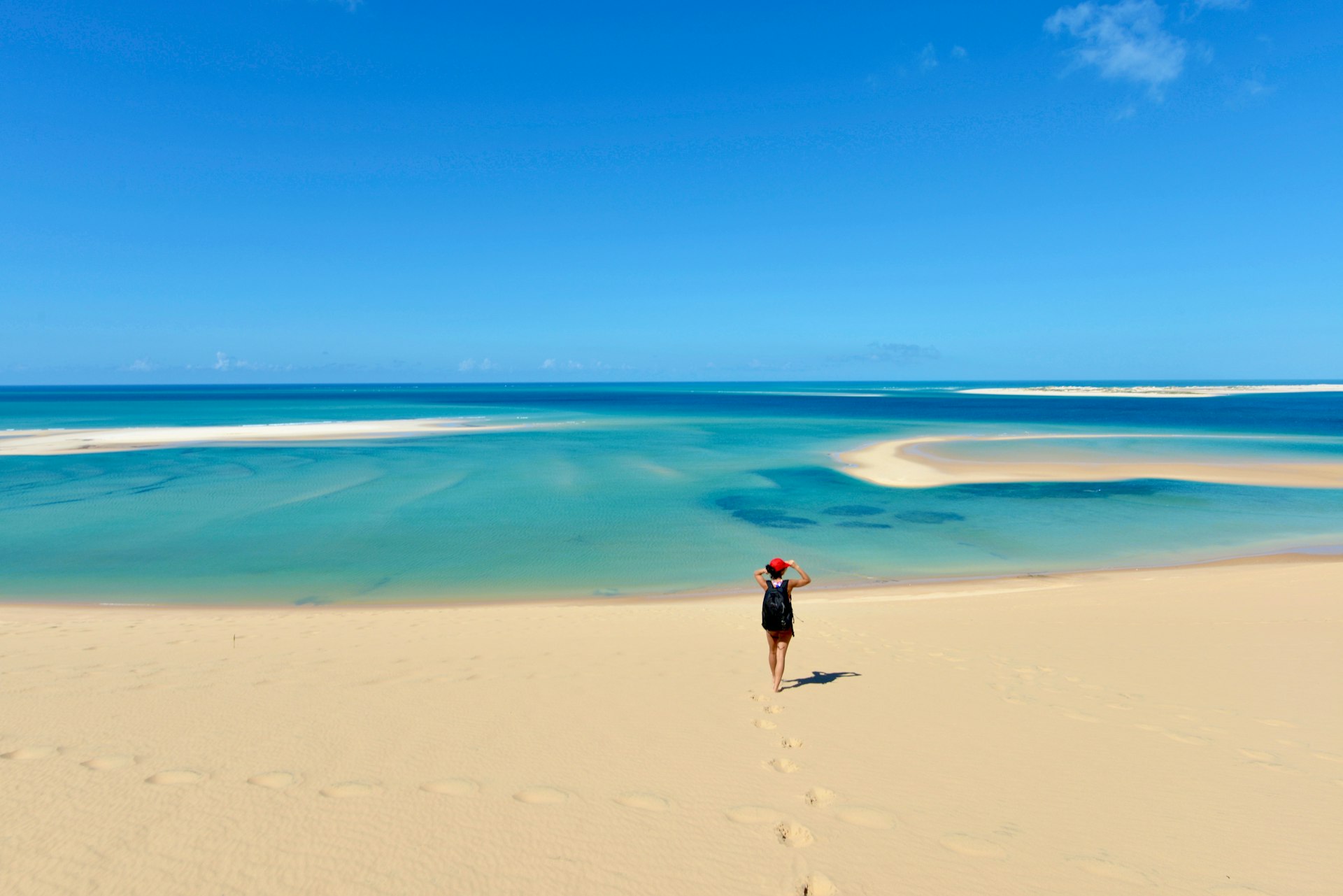 A lone traveler standing on a vast sandy beach, observing the tranquil blue-green waters merging with the sky at the horizon, with footprints trailing behind in the sand.