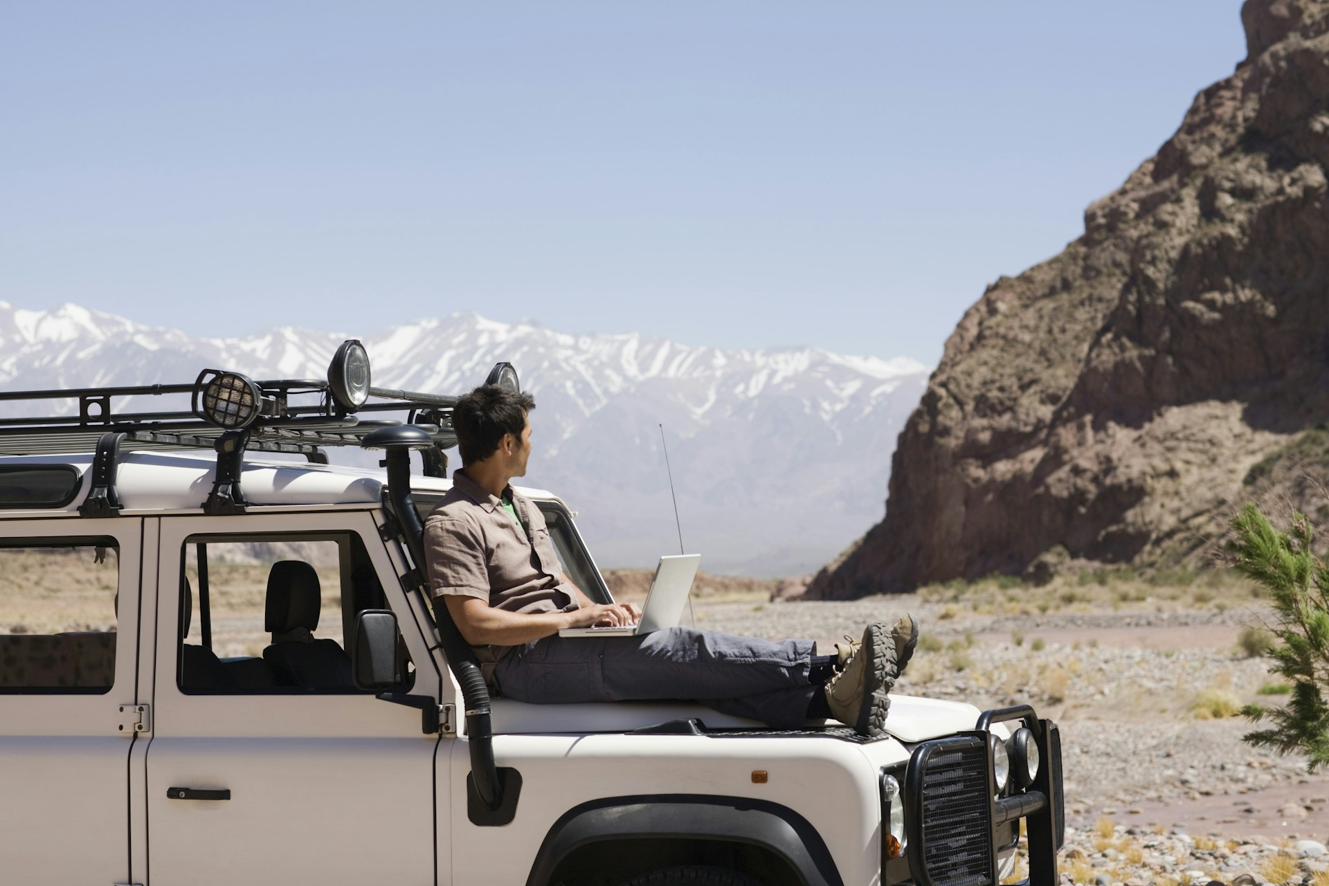 A person is perched on the hood of an off-road vehicle with a laptop, with a mountainous desert landscape in the background.