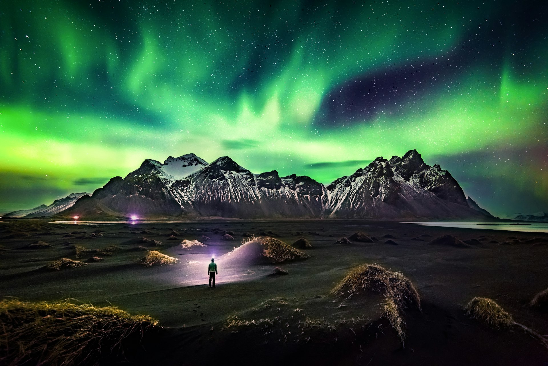A lone figure stands on a black sandy beach at night, illuminated by a headlamp, with the majestic Northern Lights casting a green glow over the rugged mountain peaks in the background.