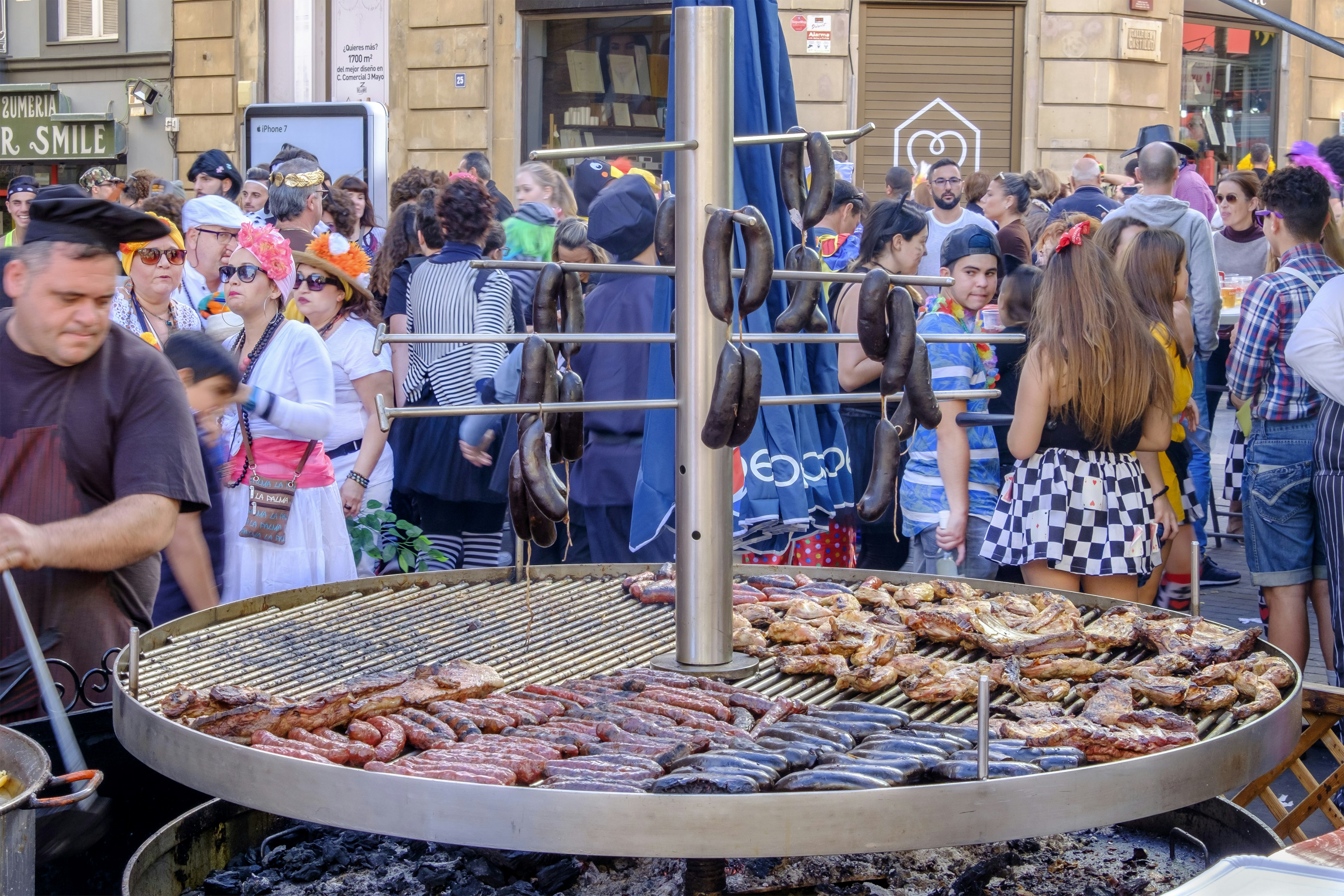 A bustling street food market with a large grill cooking various meats, surrounded by a crowd of diverse people, including some in colorful costume