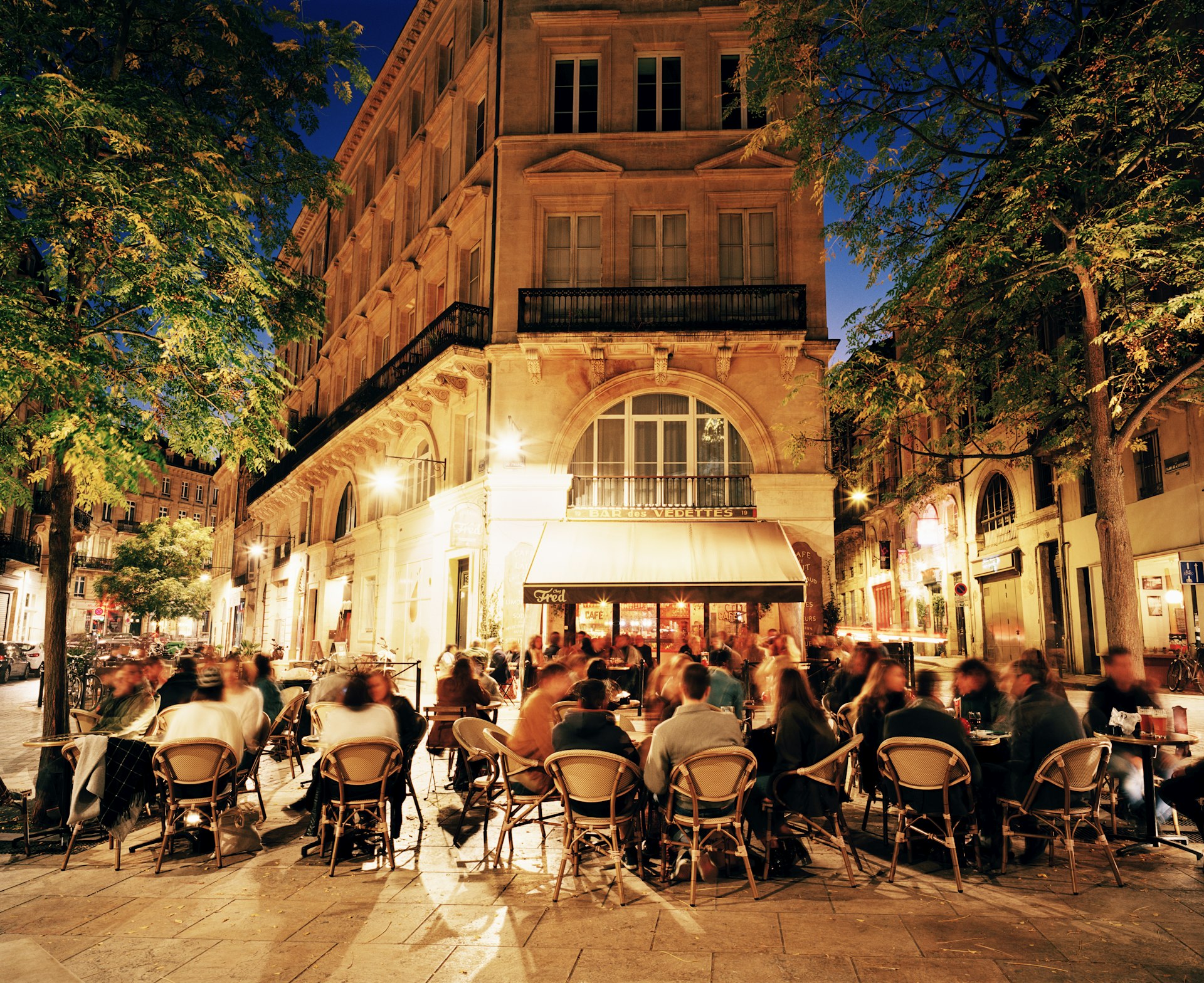 An outdoor cafe scene at night with patrons seated at tables under the glow of street lamps and a warmly lit building façade, creating a lively evening atmosphere