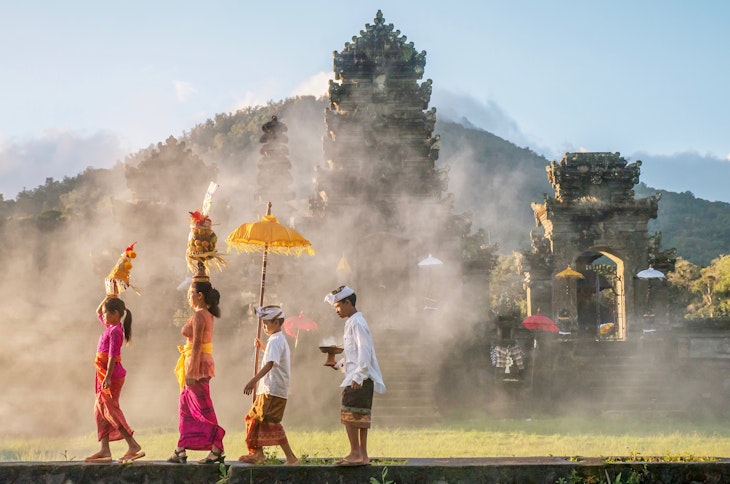 A mother and a teenaged girl are dressed in brightly colored sarongs, blouses, and sashes and are balancing tall fruit baskets on their heads. Two sons are dressed in sarongs and white shirts. The family is walking in front of an old stone temple building which has a smoky atmosphere.
938016384
religious