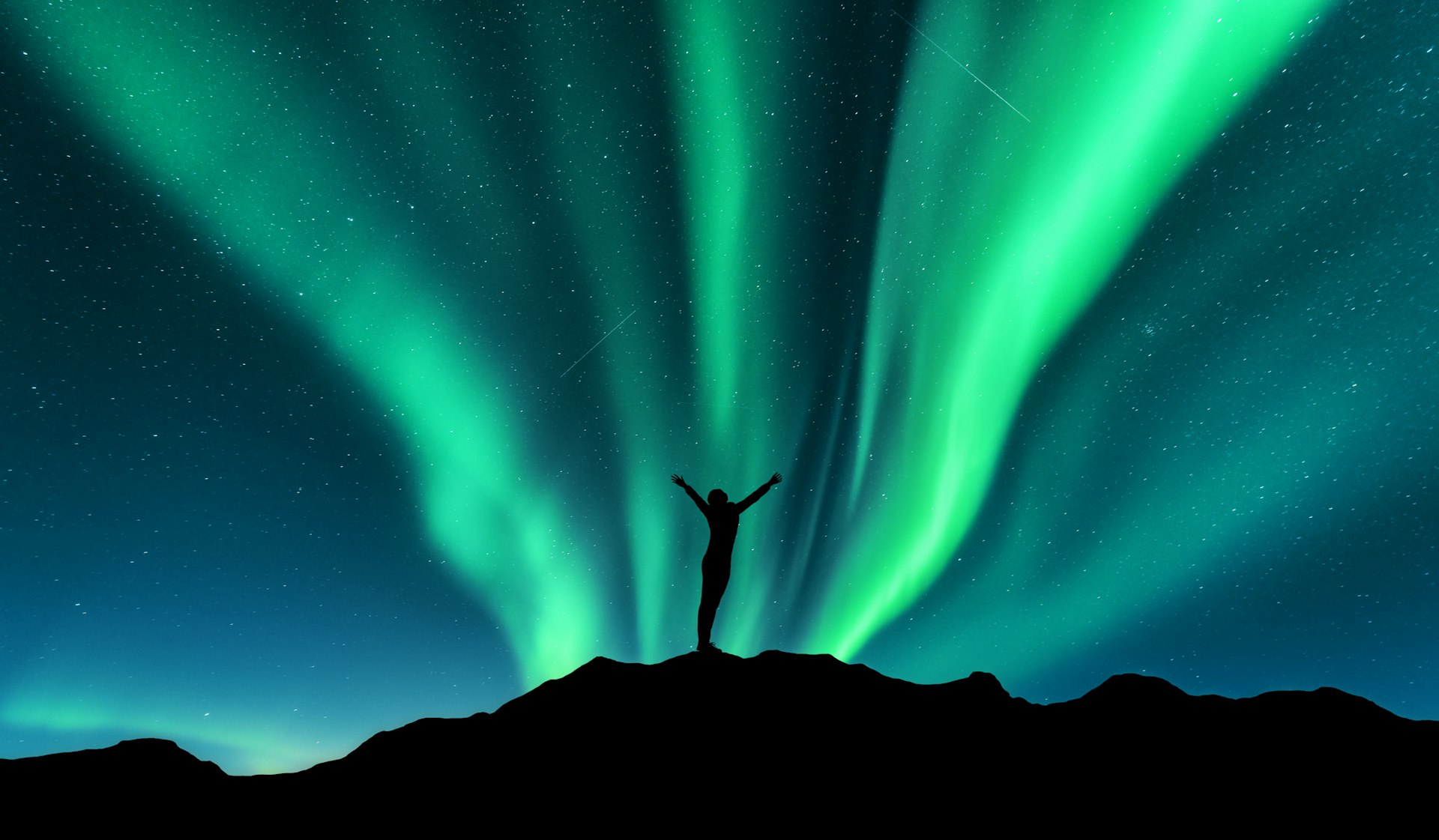 A silhouette of a person with outstretched arms standing on a hilltop against the Northern Lights, with vibrant green rays stretching across the starlit sky.