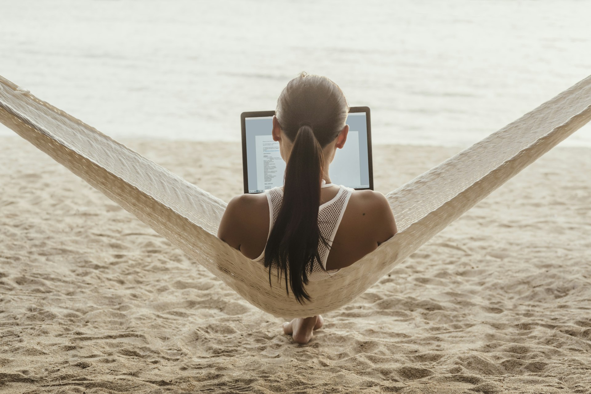 View from behind a person relaxing in a hammock with a laptop open, overlooking a sandy beach.