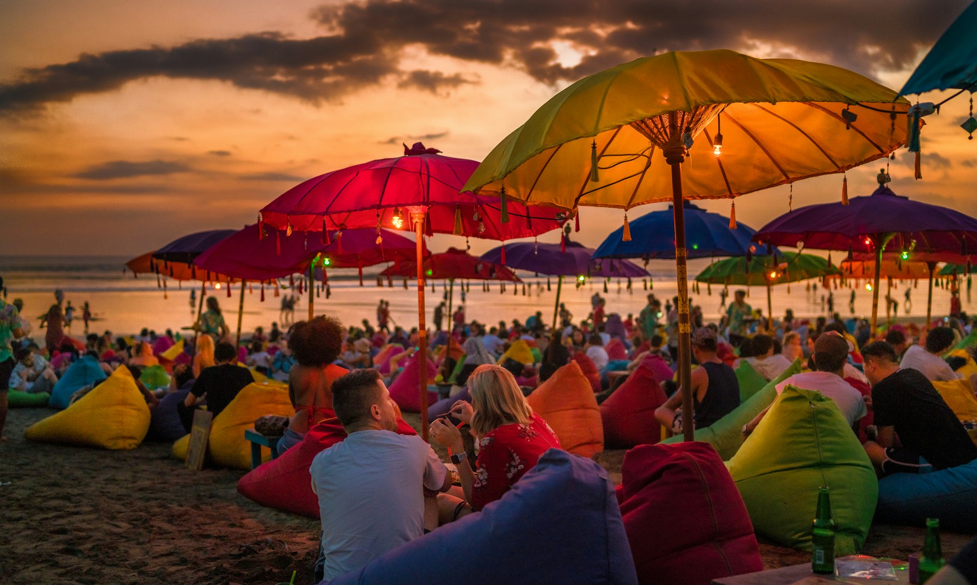 Tourists relaxing and sitting on colorful bean bags, under the umbrellas, and enjoying the sunset at Denpasar beach. Bali