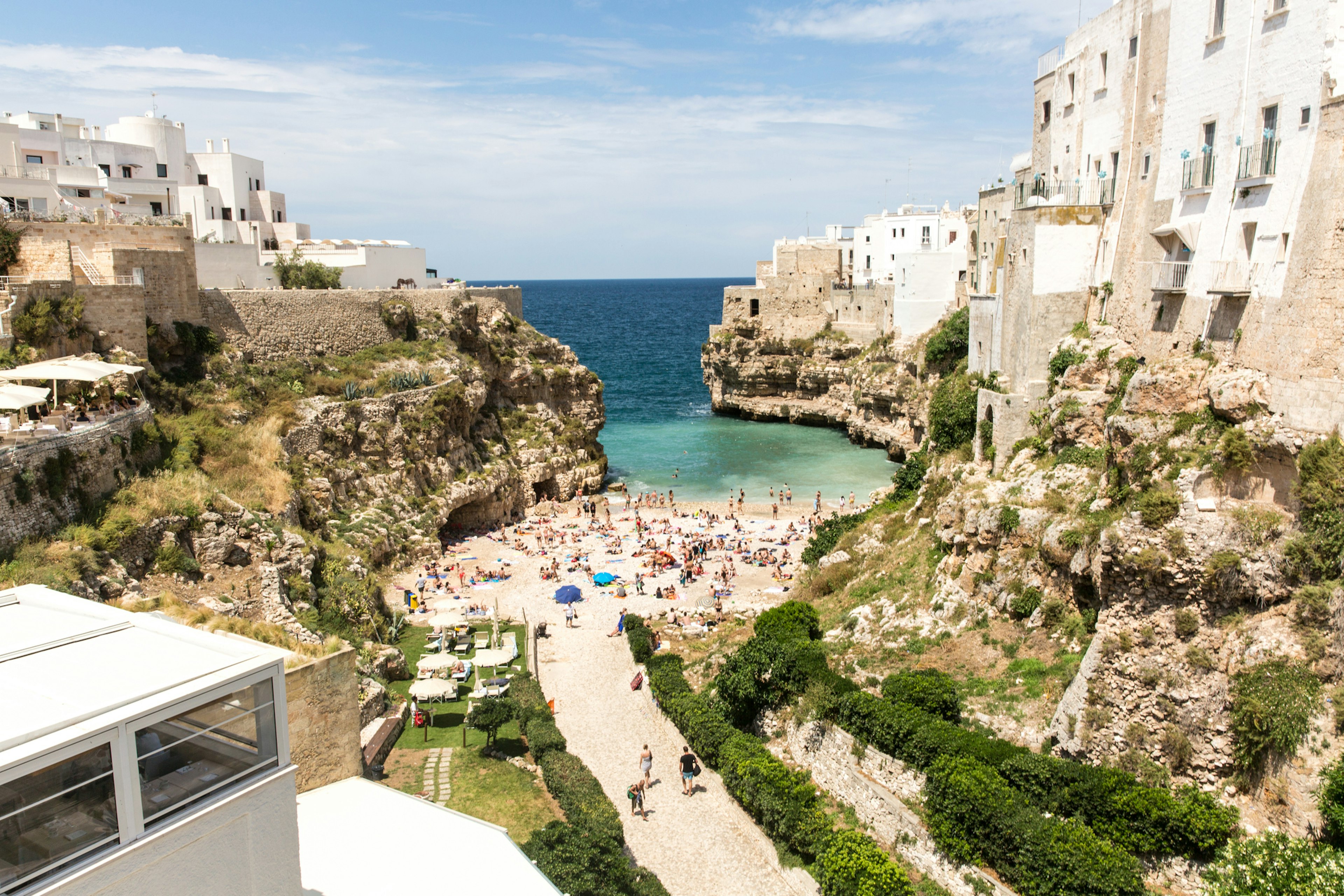 A pathway opens up to a small beach crowded with people in a beautiful cove of turquoise sea