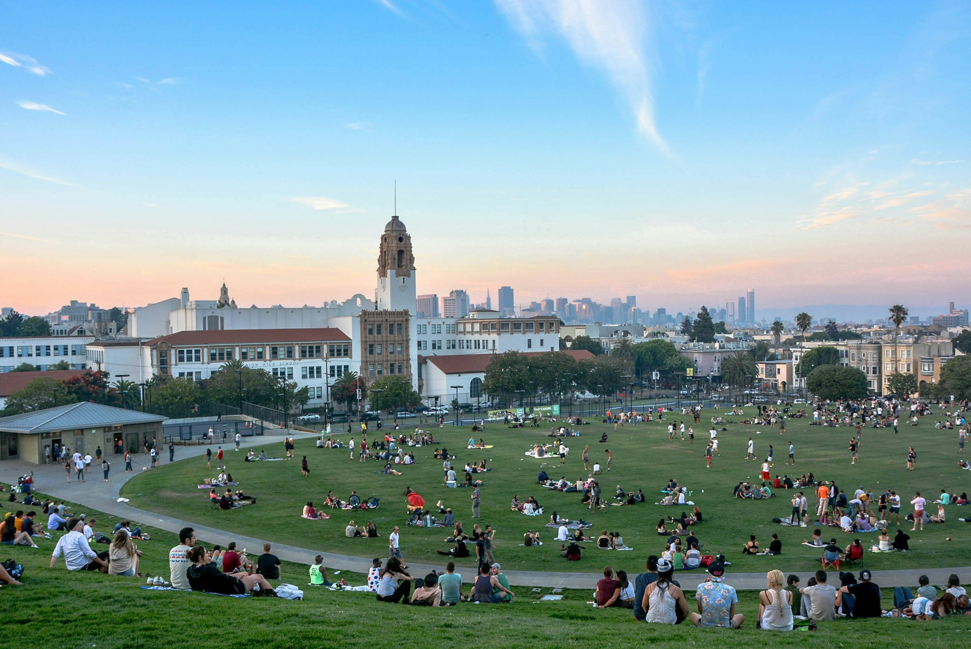 Groups of people relax on grass in a park with a city view as the sun sets