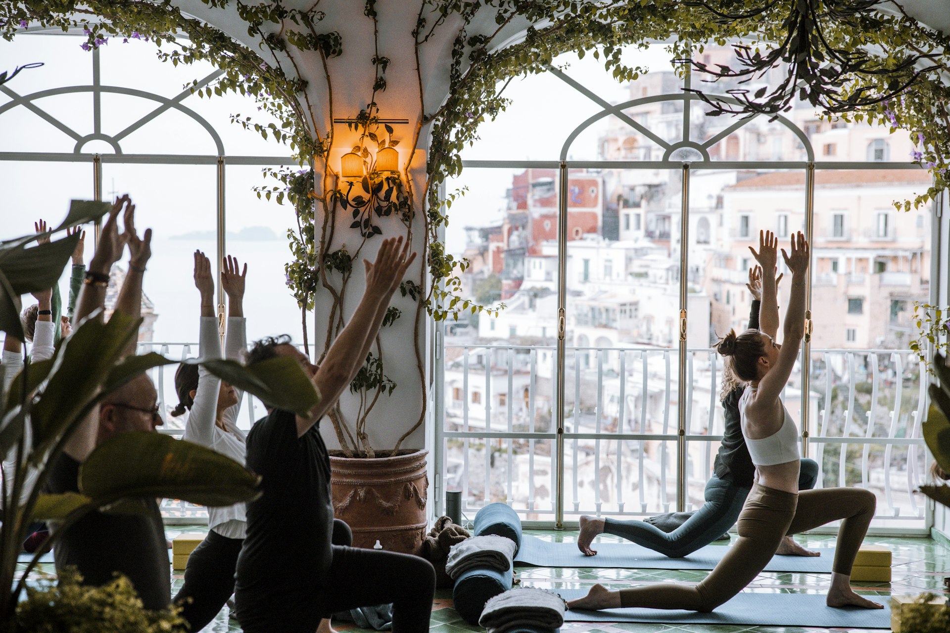 A group of people practicing yoga in a room with large windows overlooking a historic cityscape, with natural light filling the space and ornamental plants in the foreground.