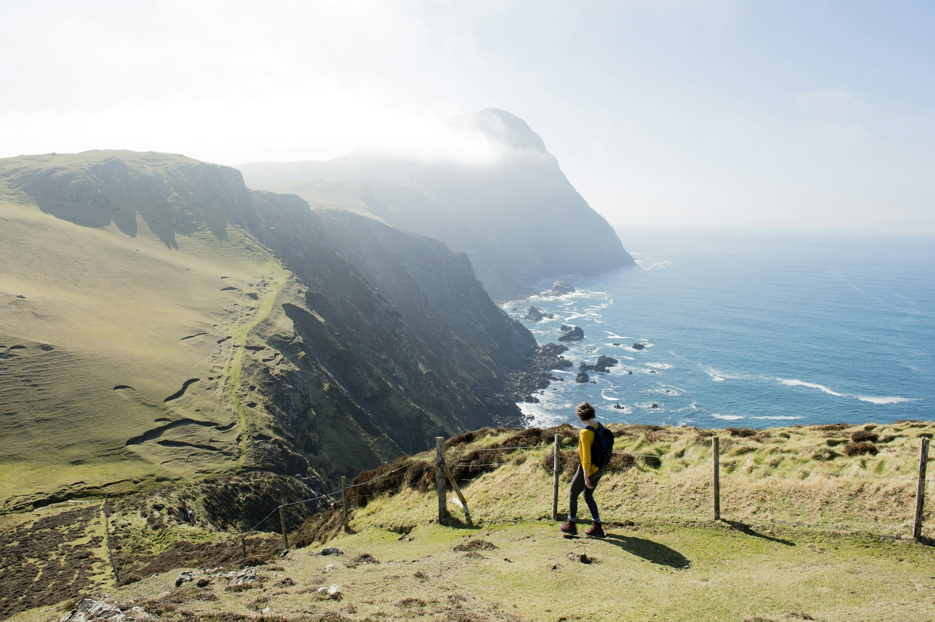 A hiker standing on a grassy cliff overlooking the ocean, with a mountain partially enveloped in mist in the background, under a clear blue sky.
