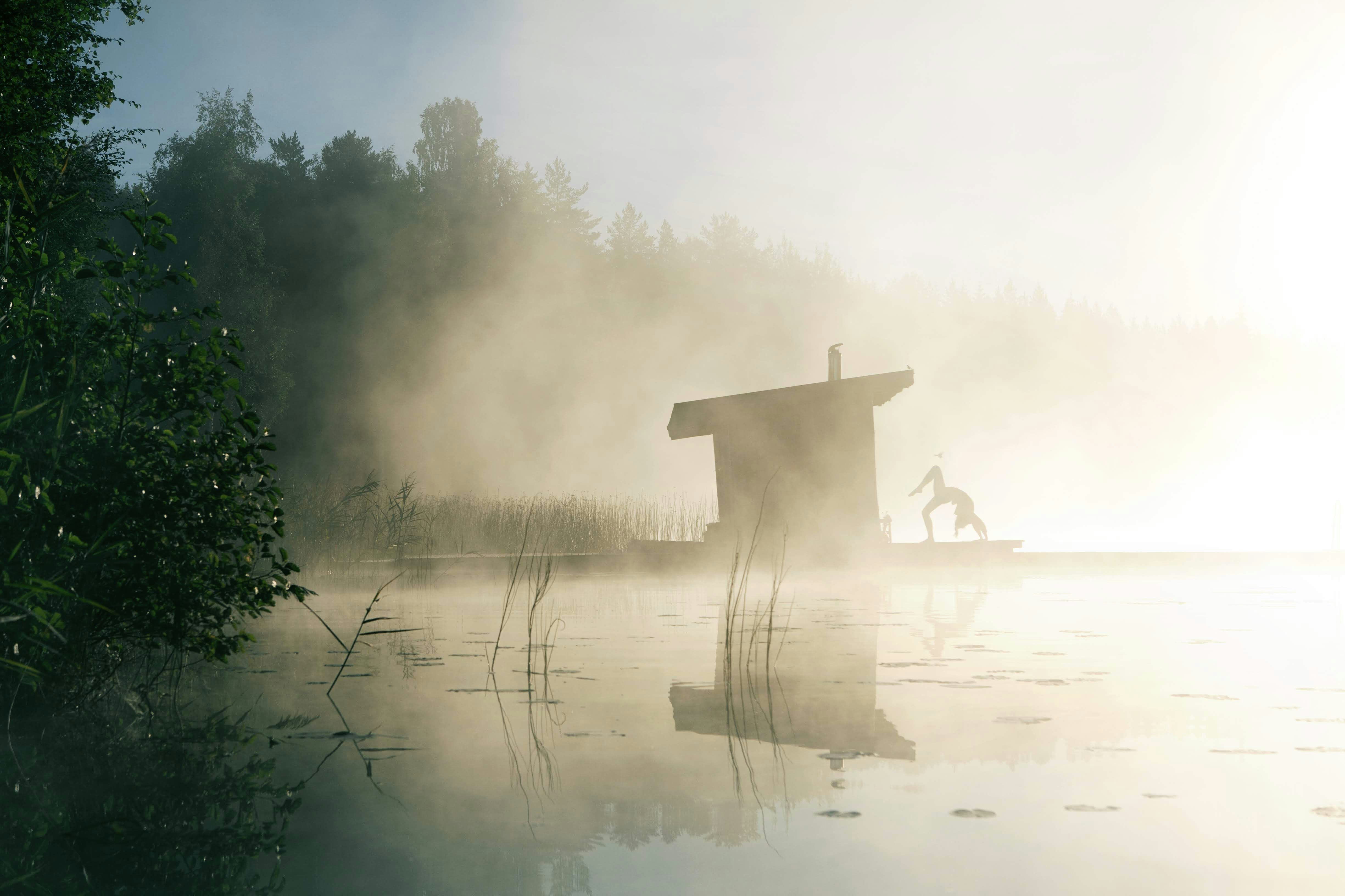 A person performing a yoga pose on a wooden dock in a misty lakeside setting, with the morning sun creating a bright backdrop and the surrounding nature partially obscured by fog