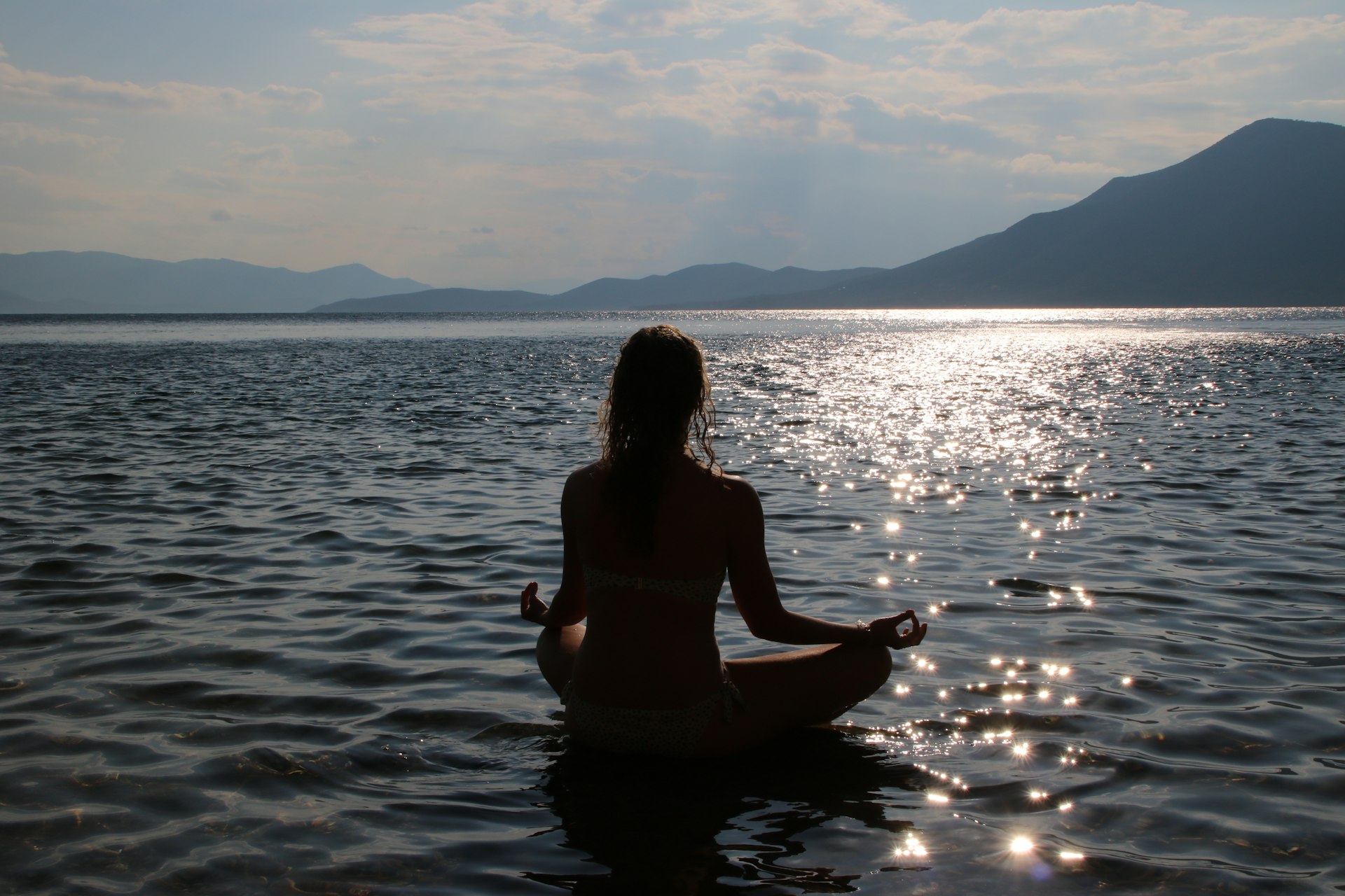 A silhouette of a person meditating in the lotus position by the sea at sunset, with mountains in the background and the water reflecting the sunlight