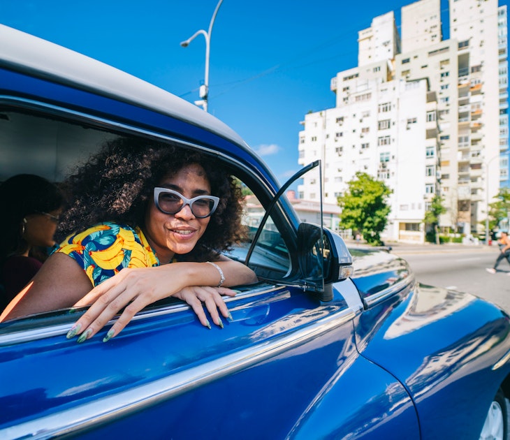 A beautiful Cuban woman with afro hair, smiles looking at the camera from inside an old blue taxi typical of Havana in Cuba on a road with a building in the background
A beautiful Cuban woman with afro hair, smiles looking at the camera from inside an old blue taxi typical of Havana in Cuba on a road with a building in the background