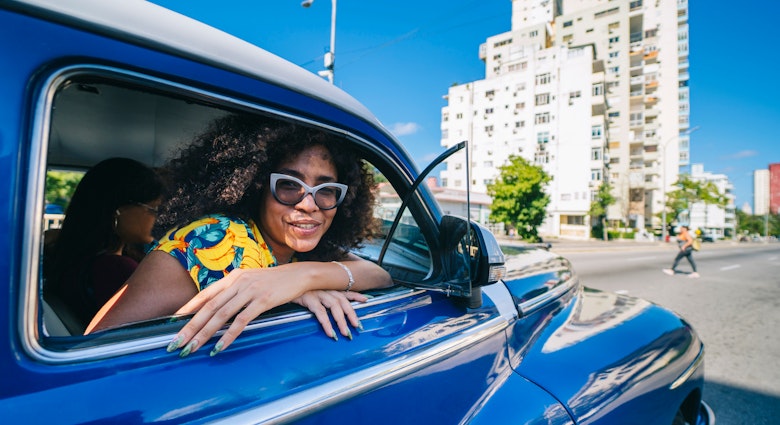 A beautiful Cuban woman with afro hair, smiles looking at the camera from inside an old blue taxi typical of Havana in Cuba on a road with a building in the background
A beautiful Cuban woman with afro hair, smiles looking at the camera from inside an old blue taxi typical of Havana in Cuba on a road with a building in the background