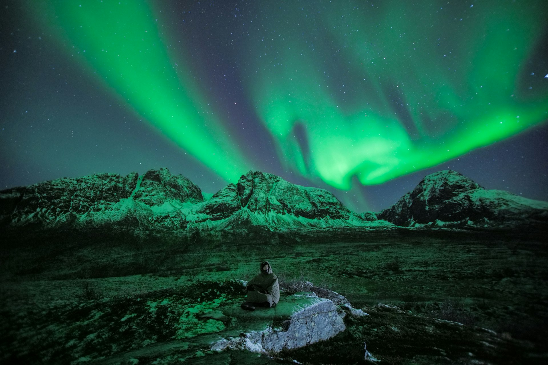 A person sits on a rock in the dark wilderness, looking up at the ethereal green glow of the Northern Lights.