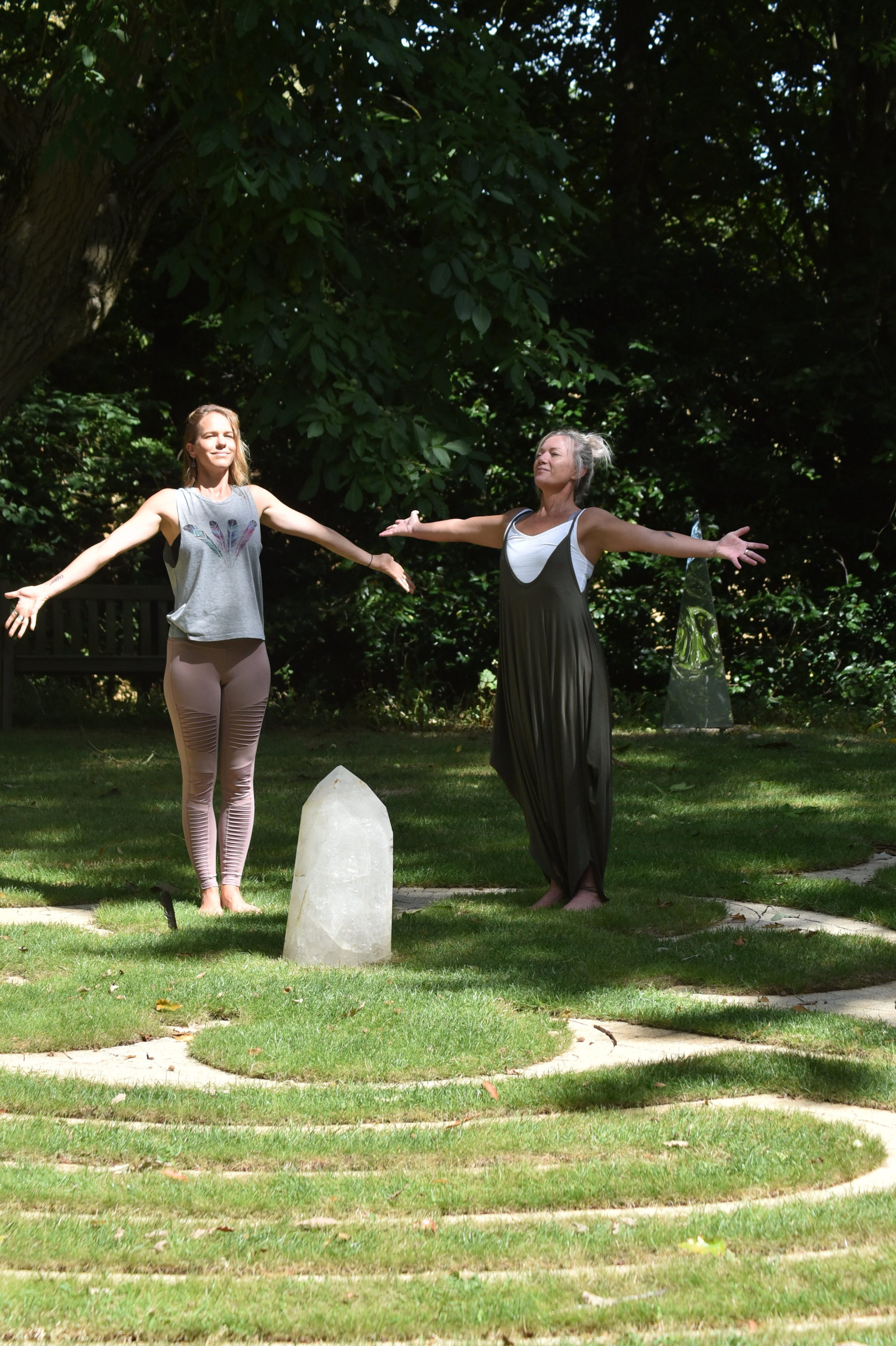 Two women standing in a garden with arms wide open, facing each other with a large clear crystal on the ground between them, surrounded by green trees.