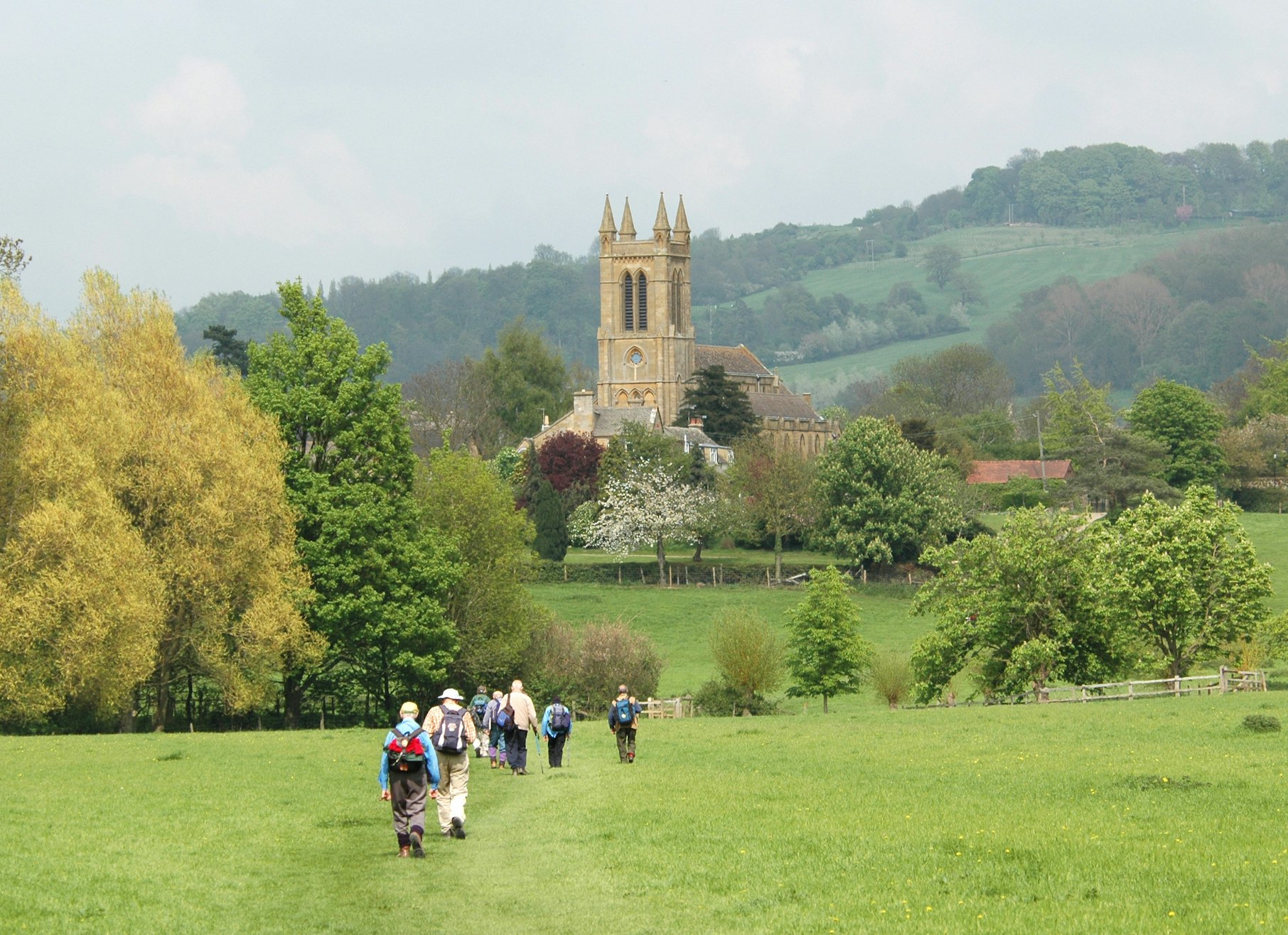 A group of walkers follow a path through a field towards a village with a church