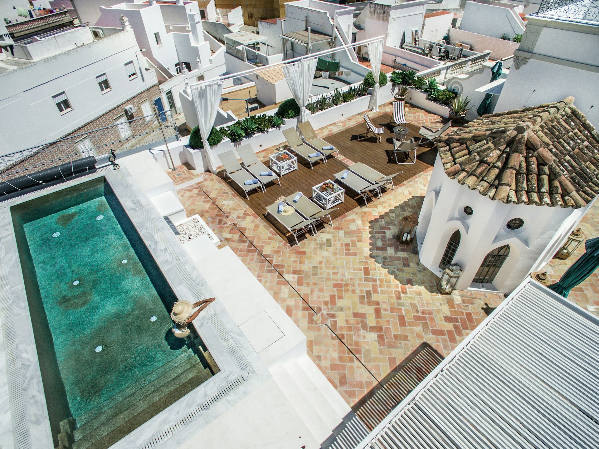 An aerial view of a rooftop terrace with a swimming pool, sun loungers and umbrellas, with the surrounding town and buildings visible in the background
