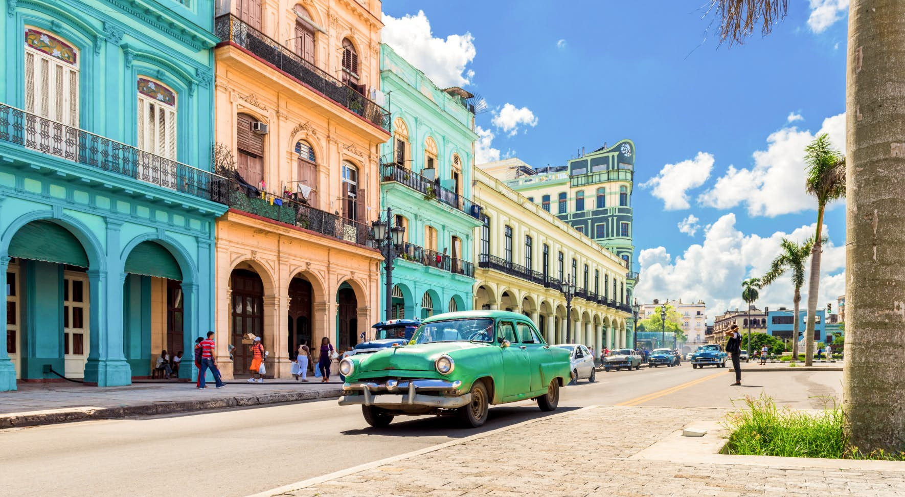 Turquoise 1950s-era car driving past brightly colored buildings