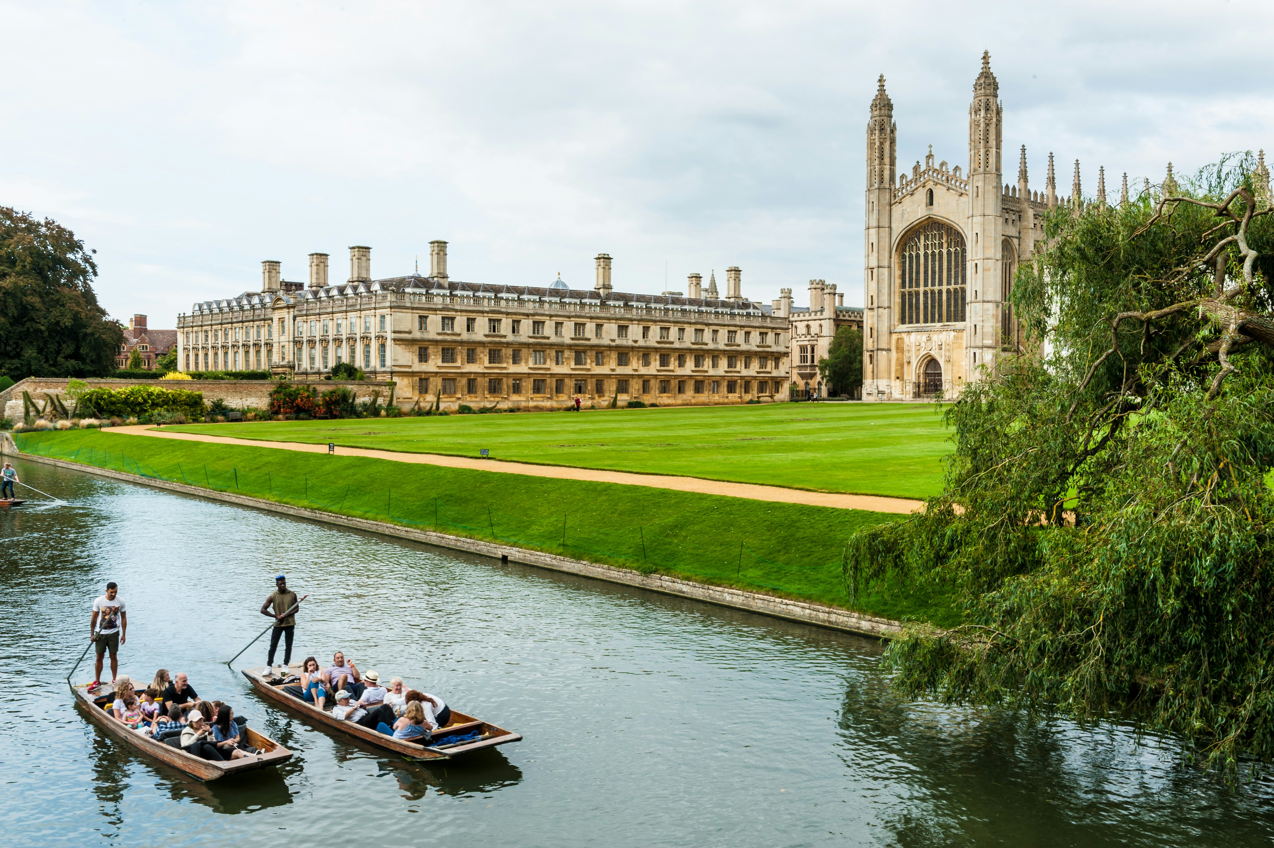 A university college and chapel near a river with two flat-bottomed boats being propelled and piloted by someone at the back using a long stick