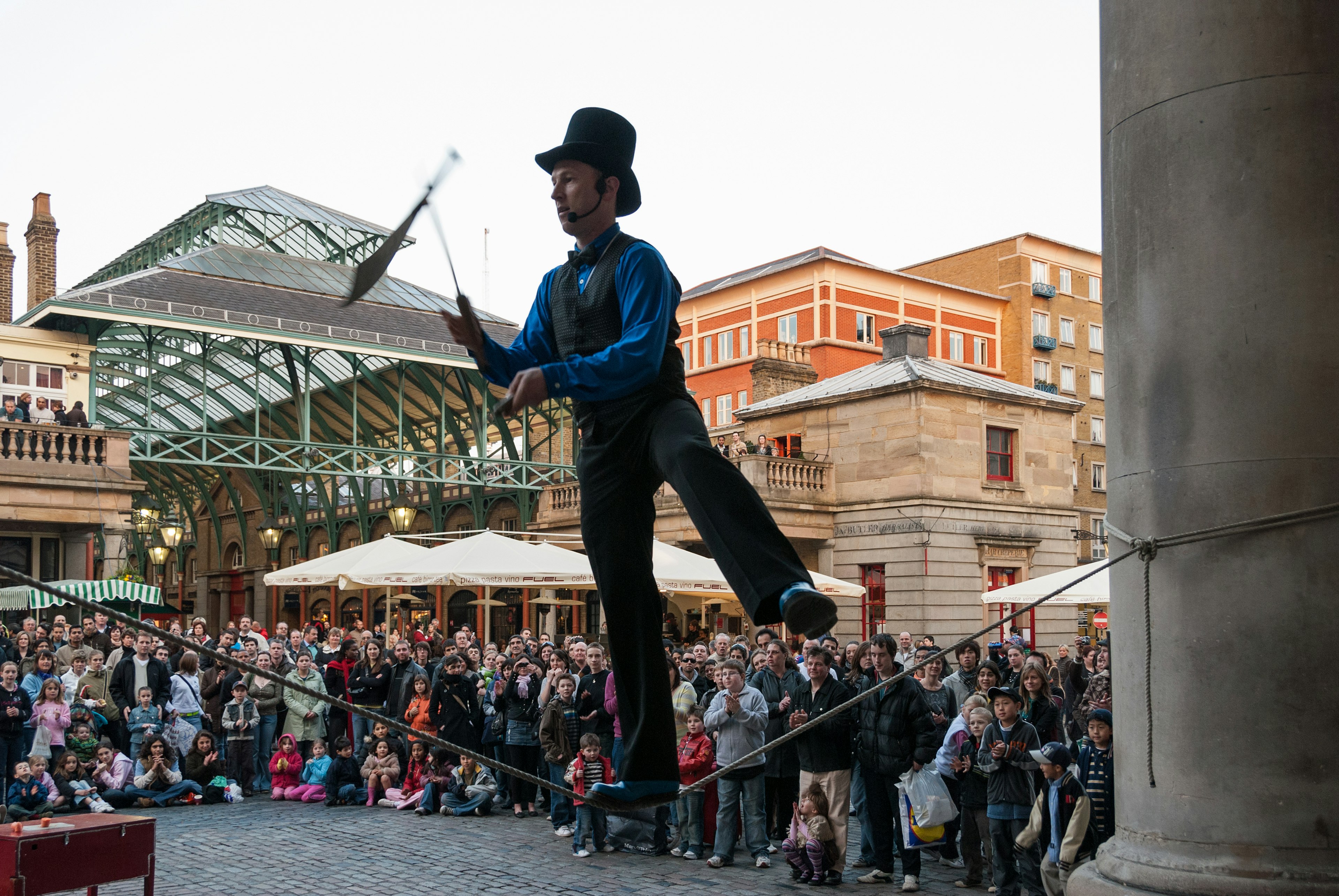 A street performer balances on a wire while juggling knives as a crowd looks on.
