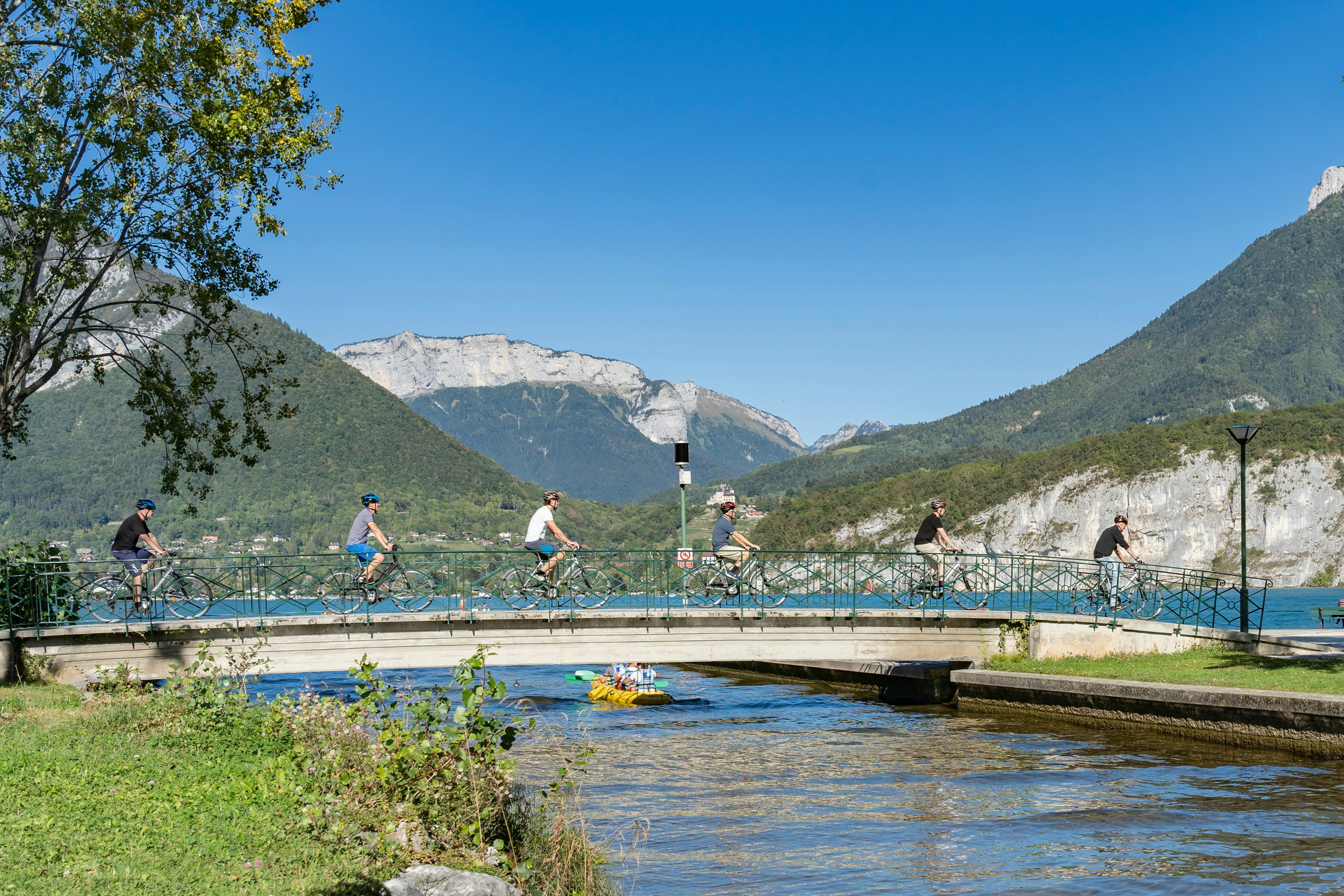 Six cyclists ride bikes over a bridge beside a lake in a mountainous region