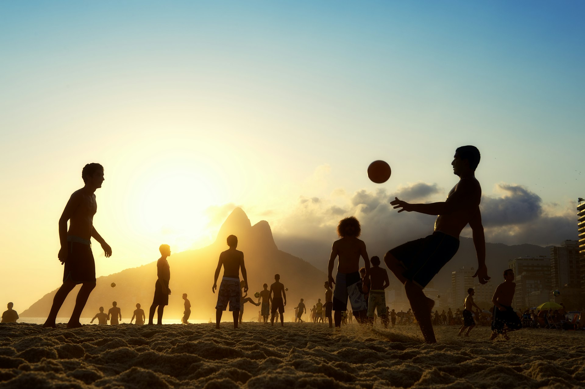 A silhouette of a group of people playing soccer on a beach at sunset, with the Dois Irmãos (Two Brothers) mountain in the background, characteristic of Rio de Janeiro's landscape.