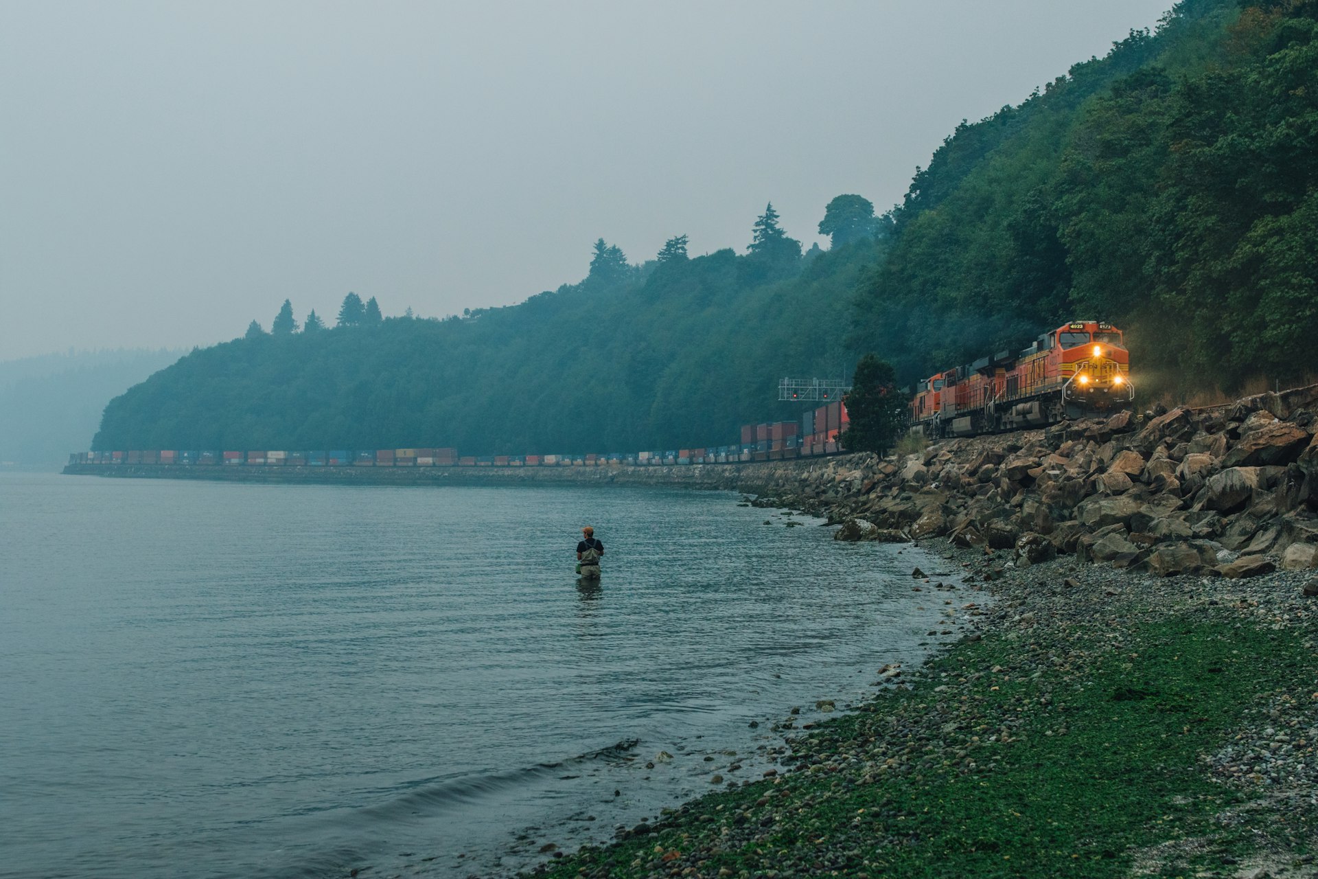 BNSF freight train traveling through Carkeek Park during a misty evening with single man fishing in Puget Sound, Seattle