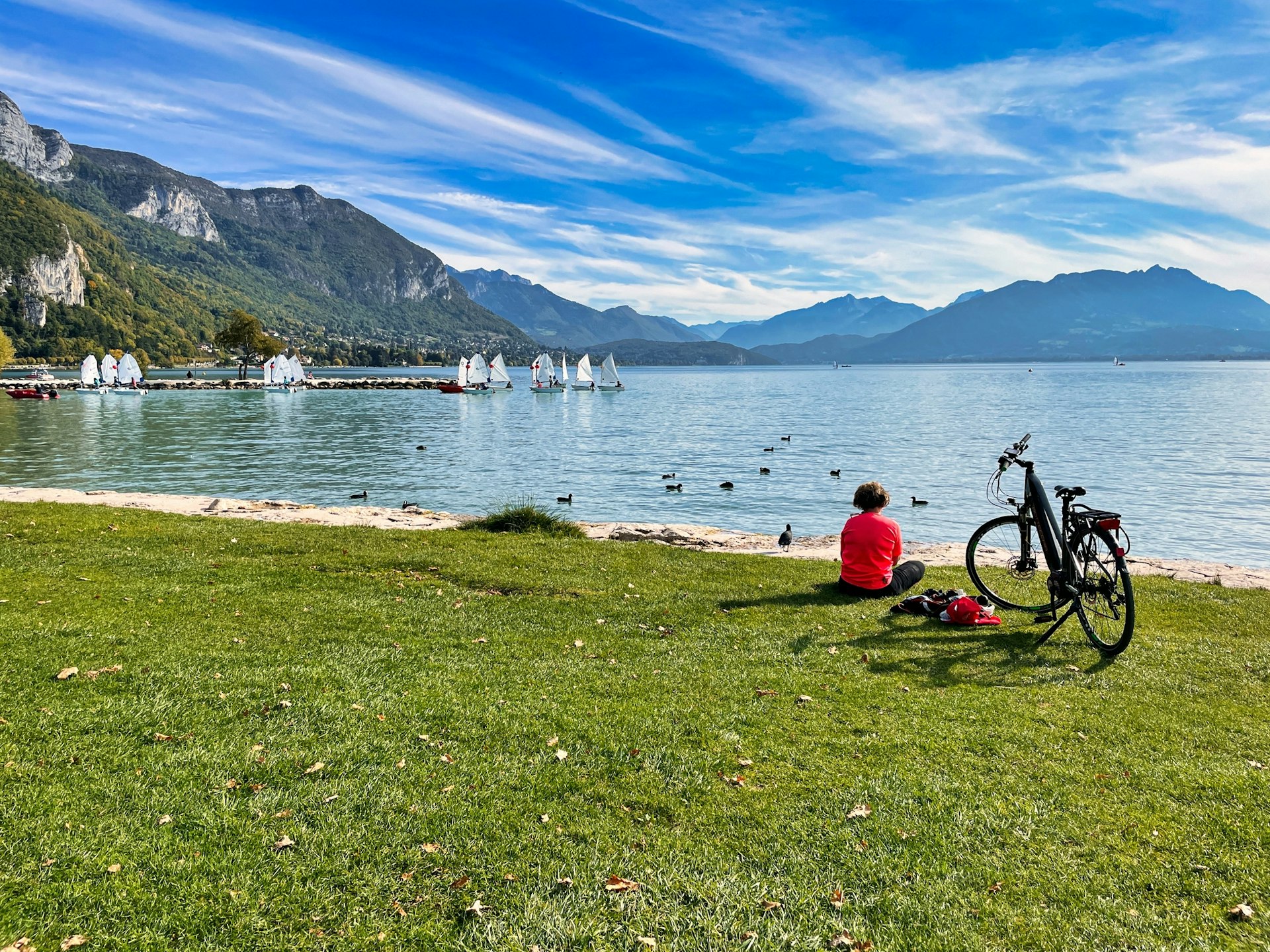 A solo cyclist sits by their bike on the lakeside enjoying the view of the sparkling waters and small sail boats in the distance