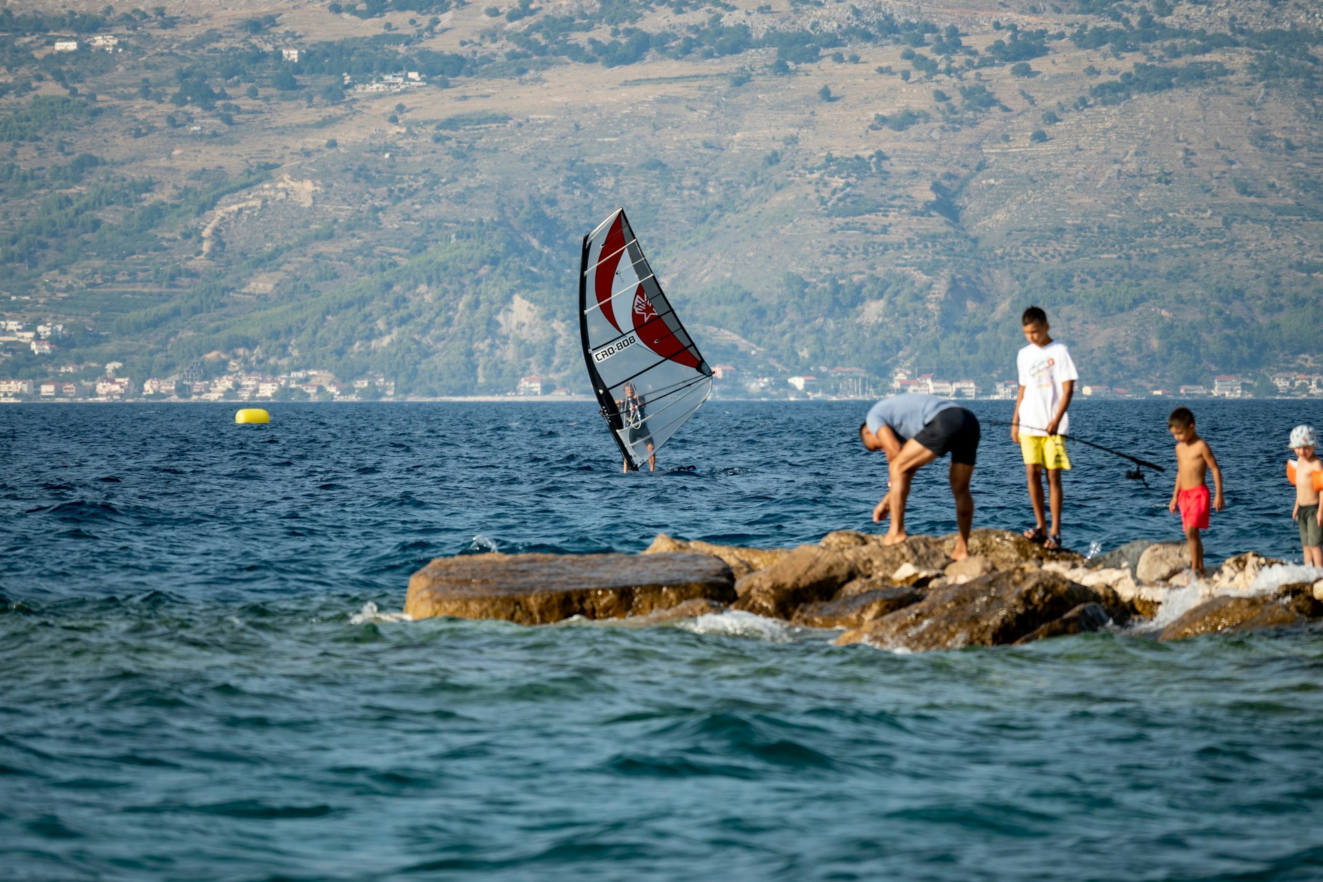 Windsurfer riding his board in front of father and his children fishing on the shore of Brac Island, Croatia