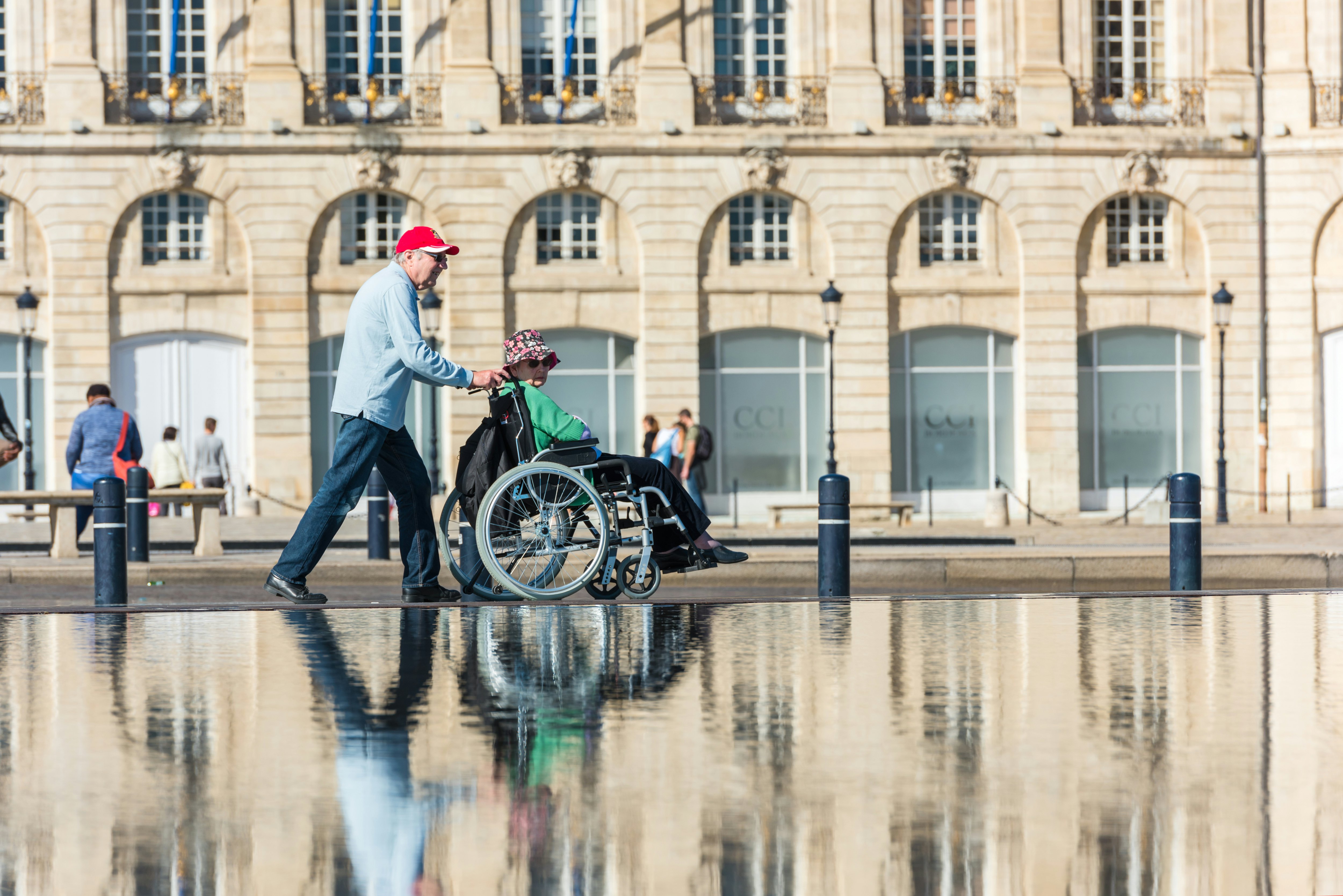 A man pushes a woman in a wheelchair in front of the Place de la Bourse, Bordeaux, Aquitaine, France