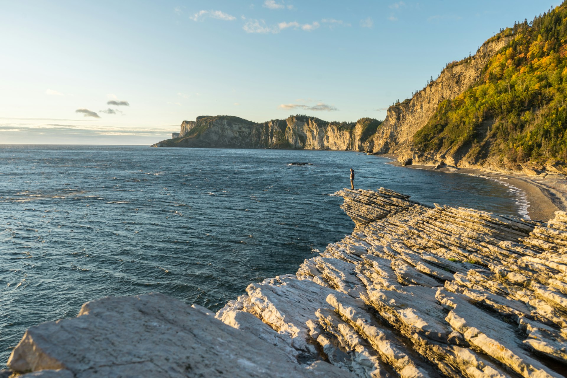 A person stands on the rocky shoreline backed by cliffs
