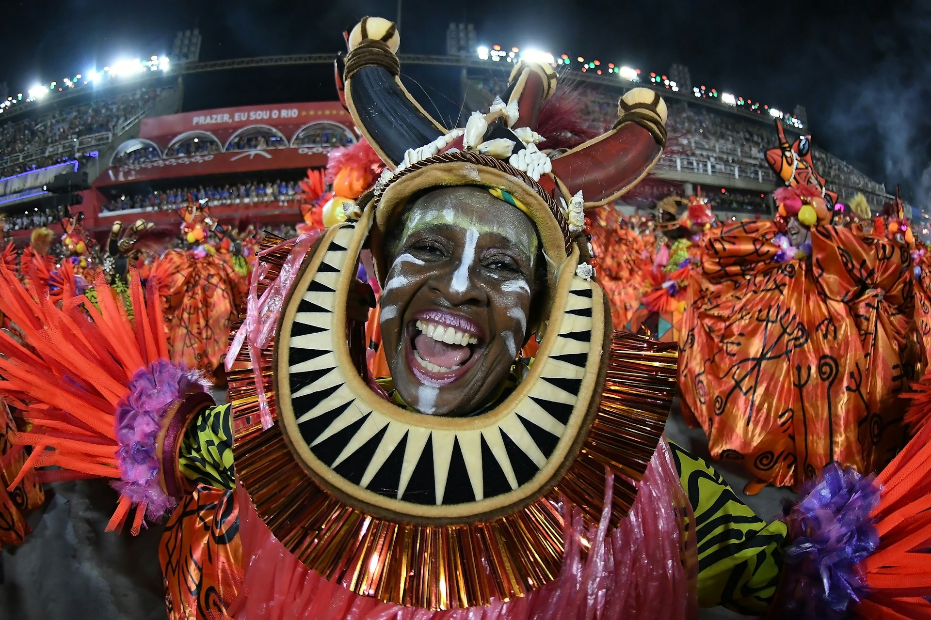 A joyous dancer at the Rio Carnival wears an elaborate tribal-themed costume with a large circular headpiece, laughing heartily during a night parade.