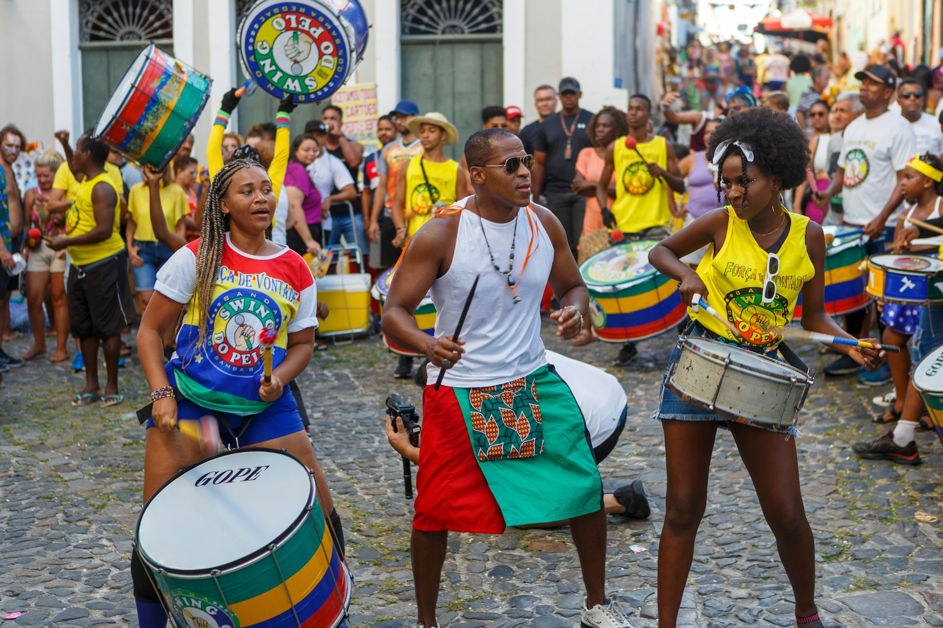 Three percussionists, two women and one man, playing drums in a street parade in Pelourinho in Salvador, Brazil with onlookers in the background in a historic area with cobblestone streets.