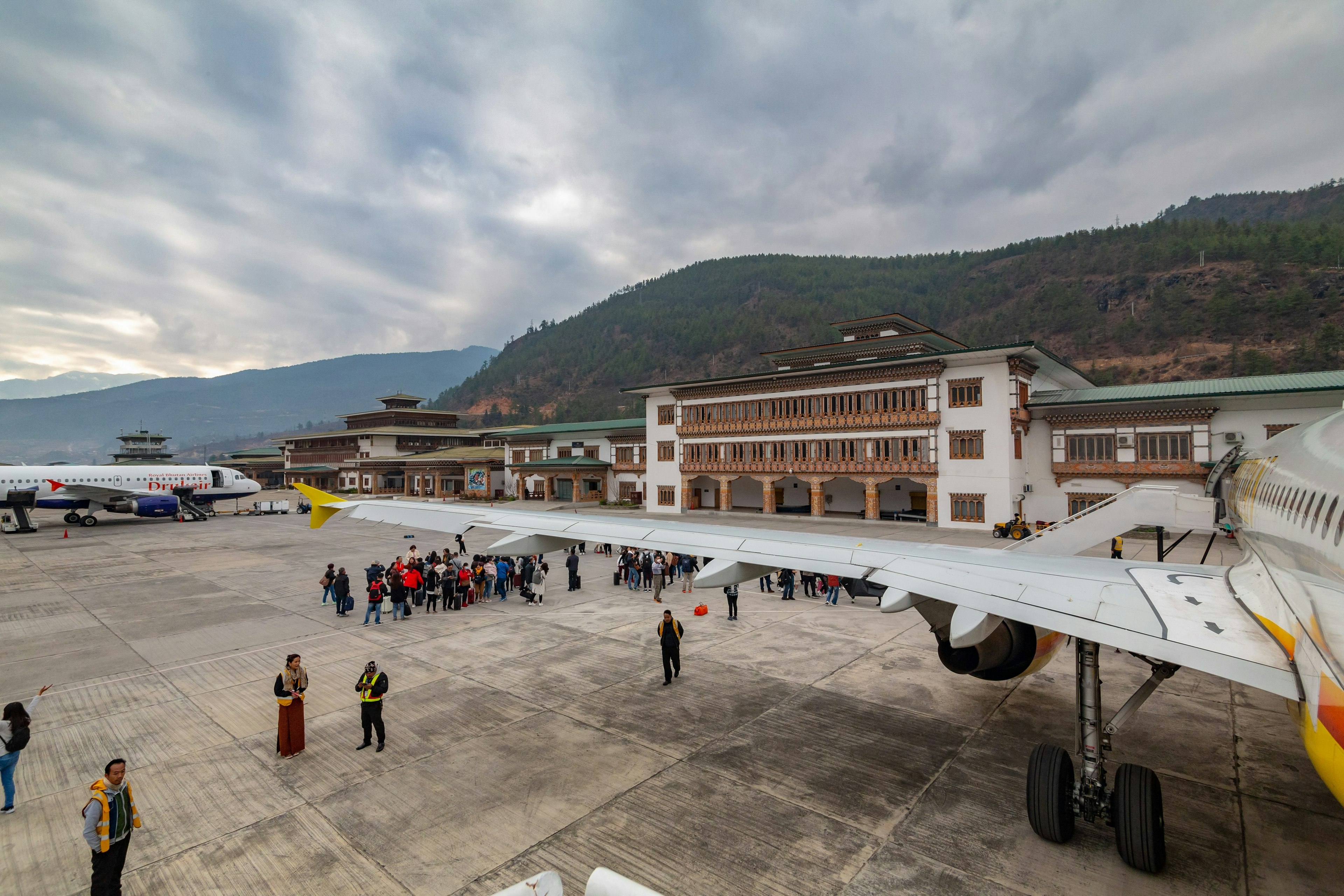 Close-up of an airplane wing with a full plane in the distance and people on the tarmac at Bhutan International Airport, seen during daytime in winter.