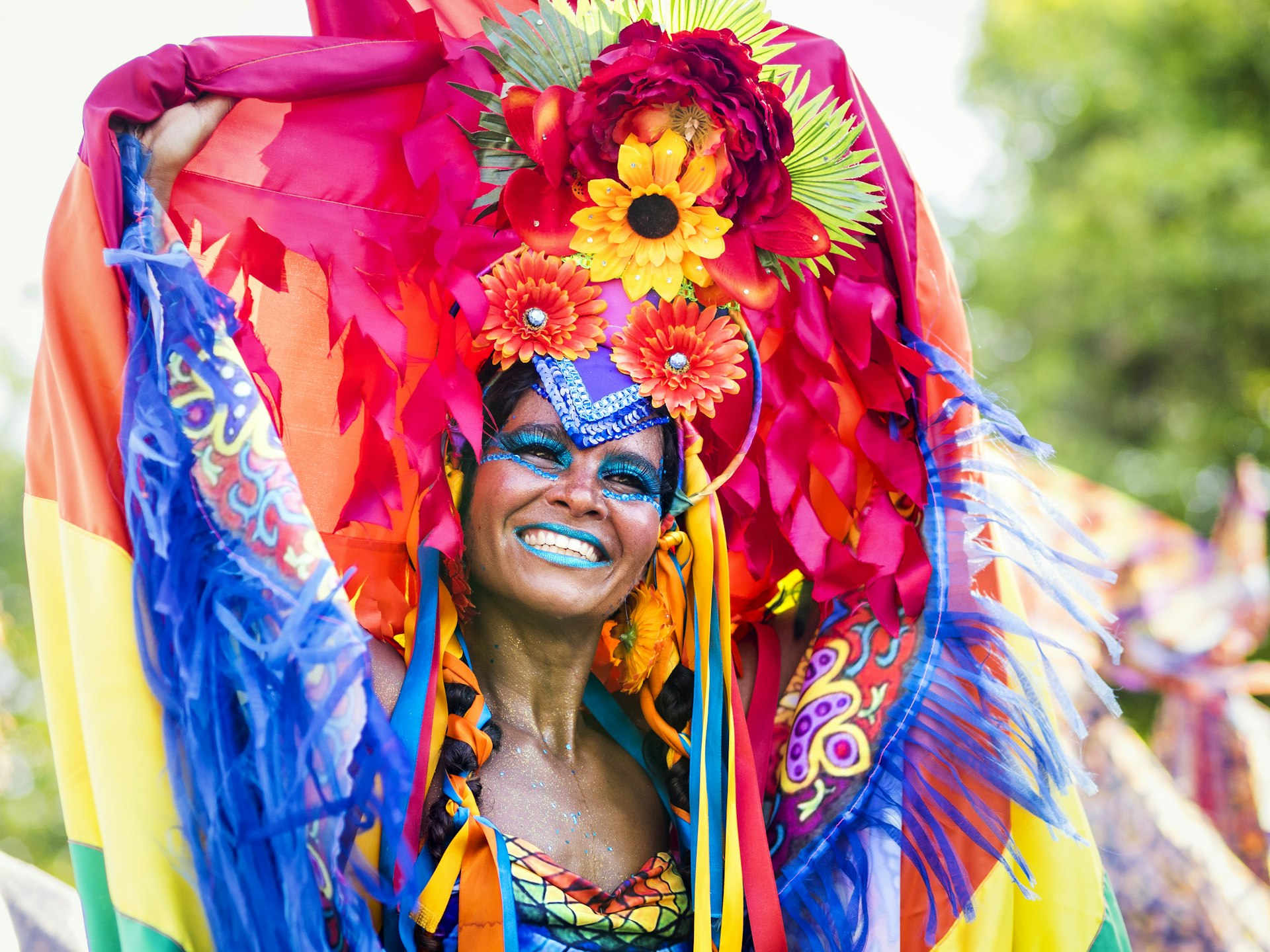 A close-up of a smiling samba dancer in a colorful costume with feathers and flowers, with blue face paint, during a bright and festive Rio Carnival celebration.
