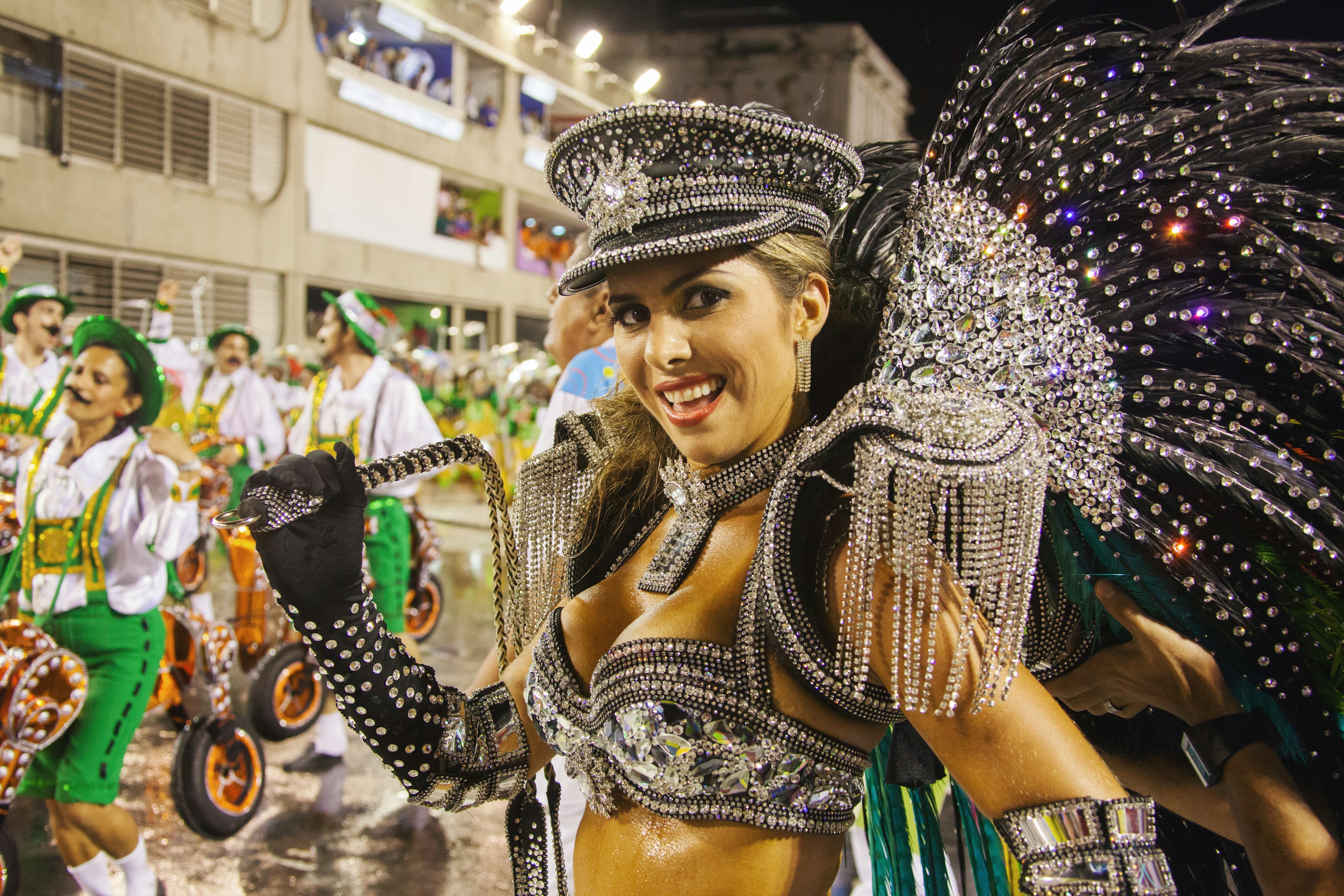 A carnival participant in a sparkling costume with black feathers and rhinestones, smiling and engaging with the camera at night during a festive parade in Rio, Brazil