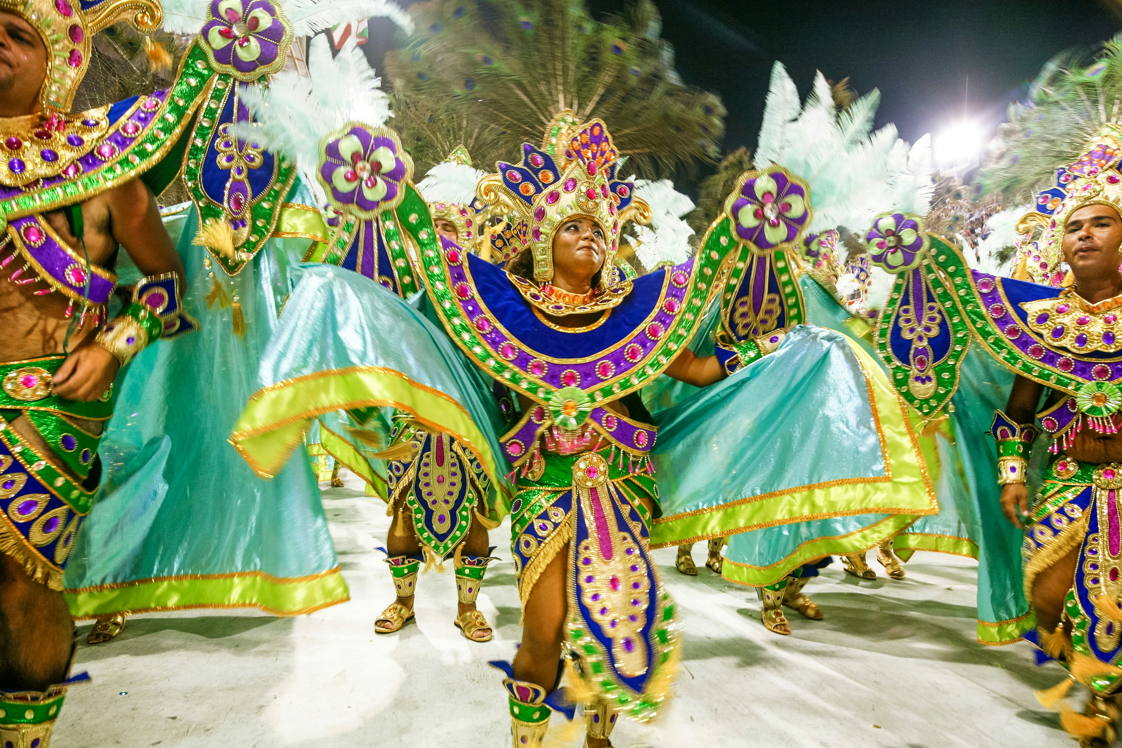 A group of dancers in vibrant, elaborate costumes with teal and purple accents, performing at a nighttime carnival event, showcasing the energy and movement of the festival.