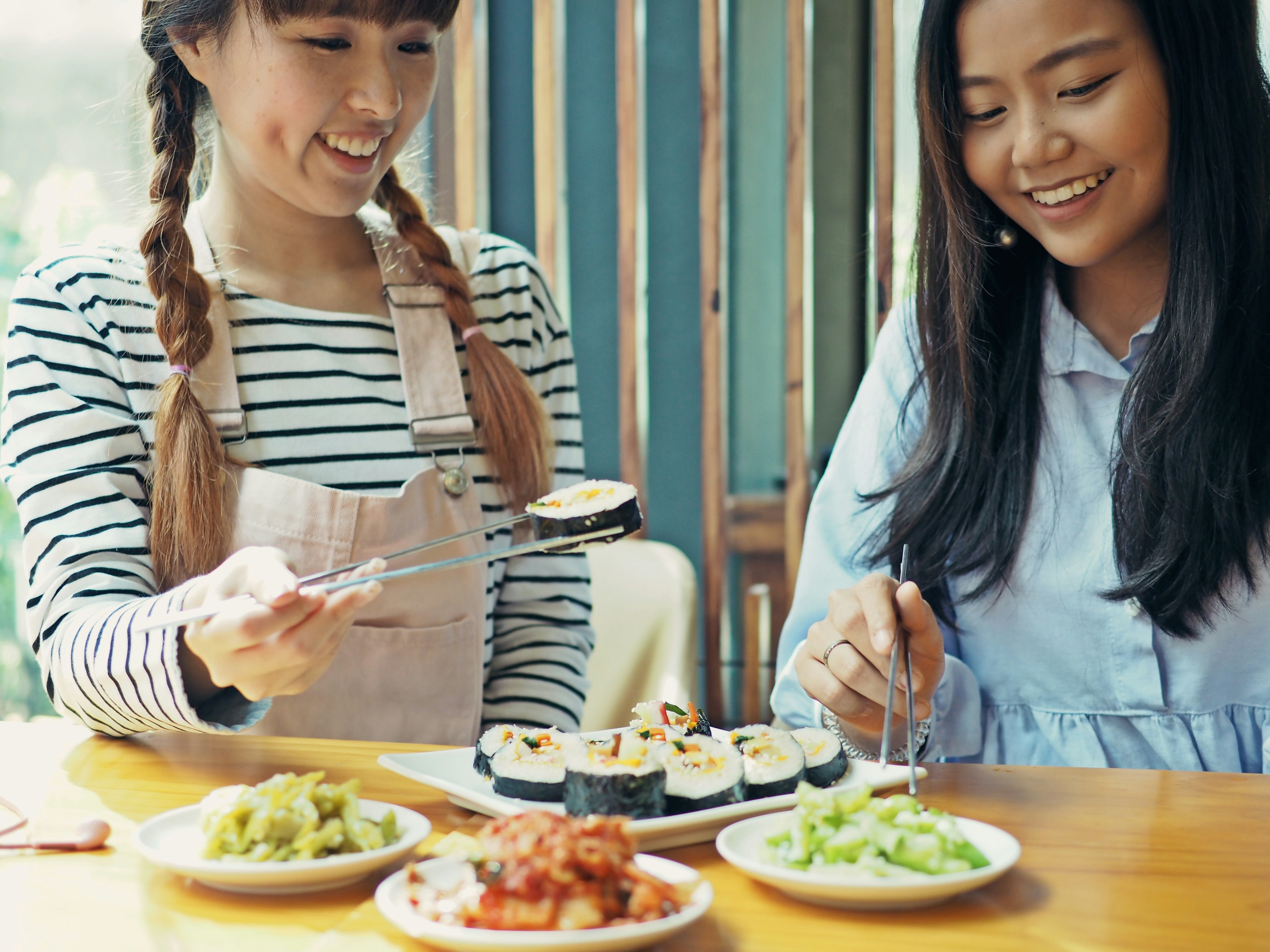 Asian women holding silver chopsticks to eat Korean seaweed rice roll with cheese or cheese kimbap (sometimes spelled gimbap) and kimchi on wooden table.