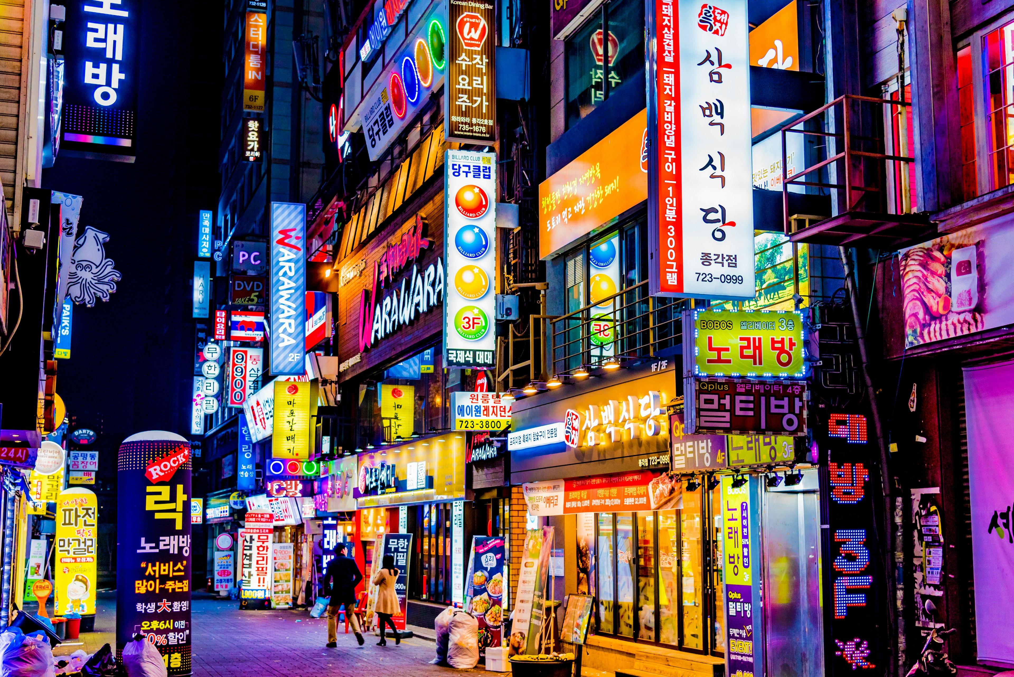 People walk down a road at night lit with bright neon signs