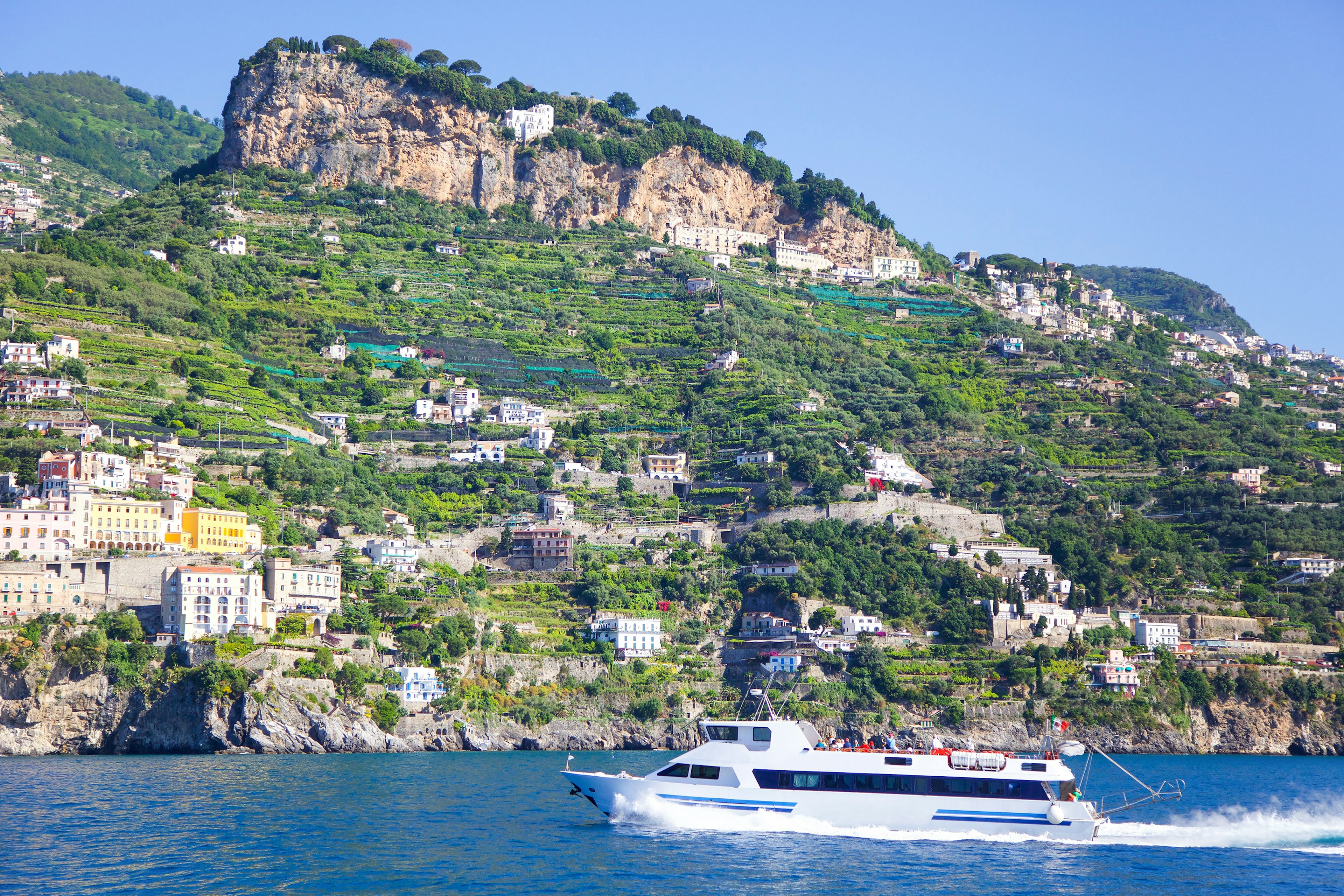 A small ferry boat transports passengers along a rocky coastline with houses built into the cliffs